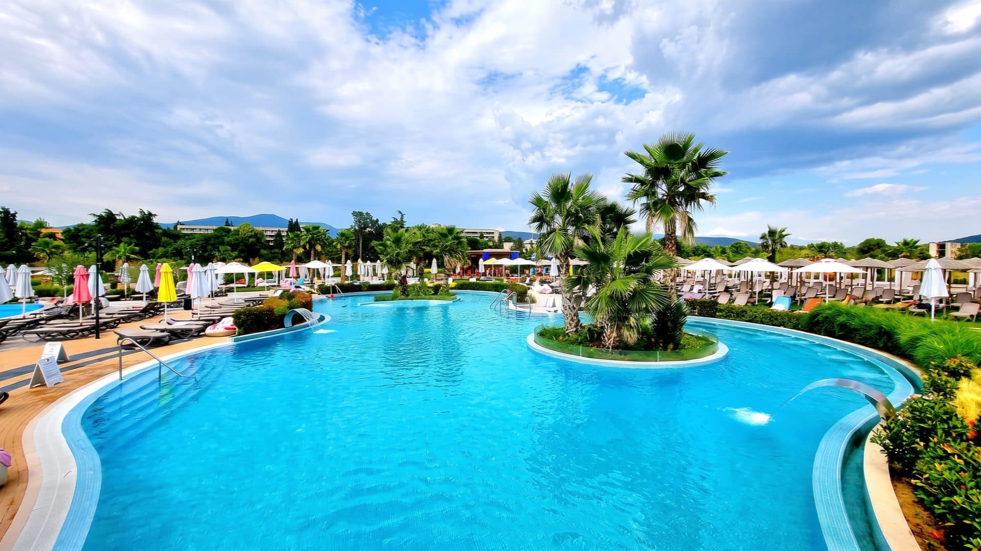 Curving blue resort pool with a palm-tree island and colourful parasols, wooded mountains rising behind.