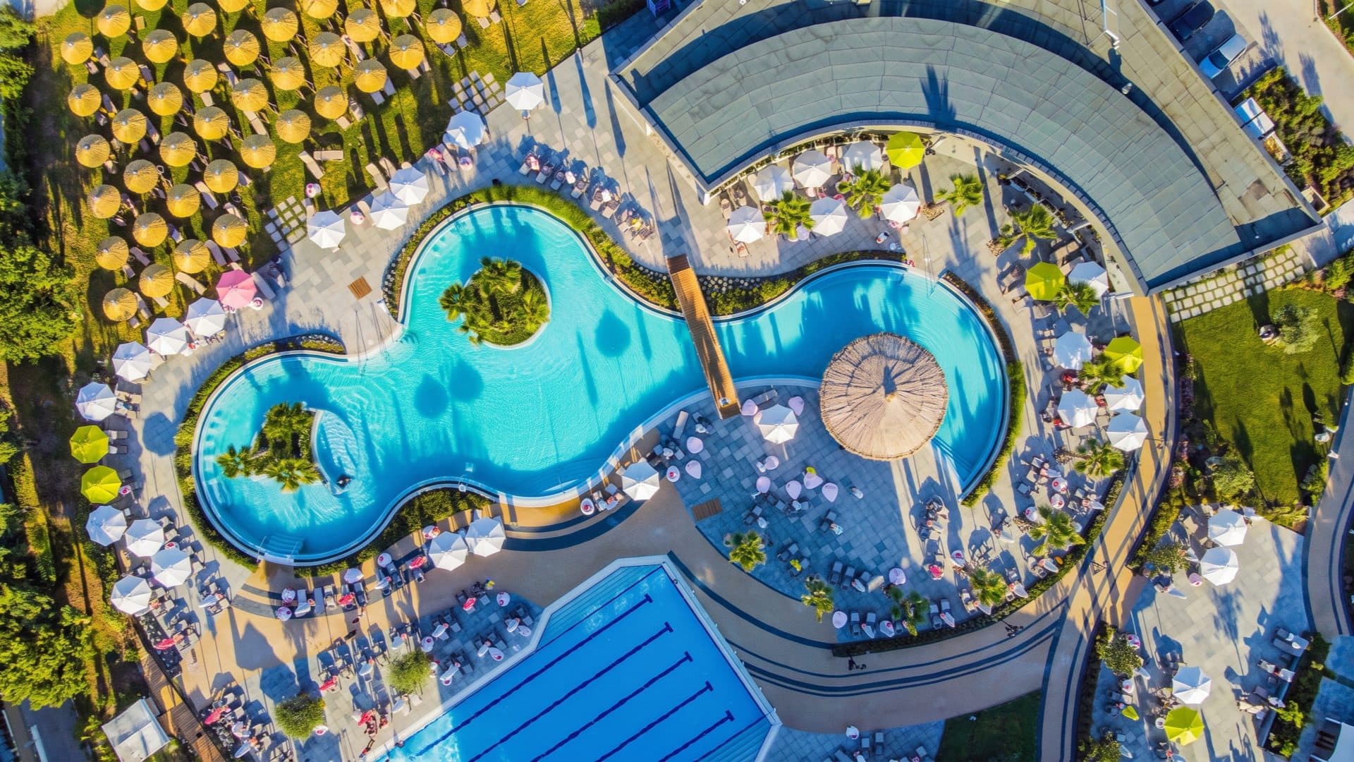 Aerial view of a figure-eight turquoise resort pool with palm islands, ringed by hundreds of straw parasols and a lap pool alongside.
