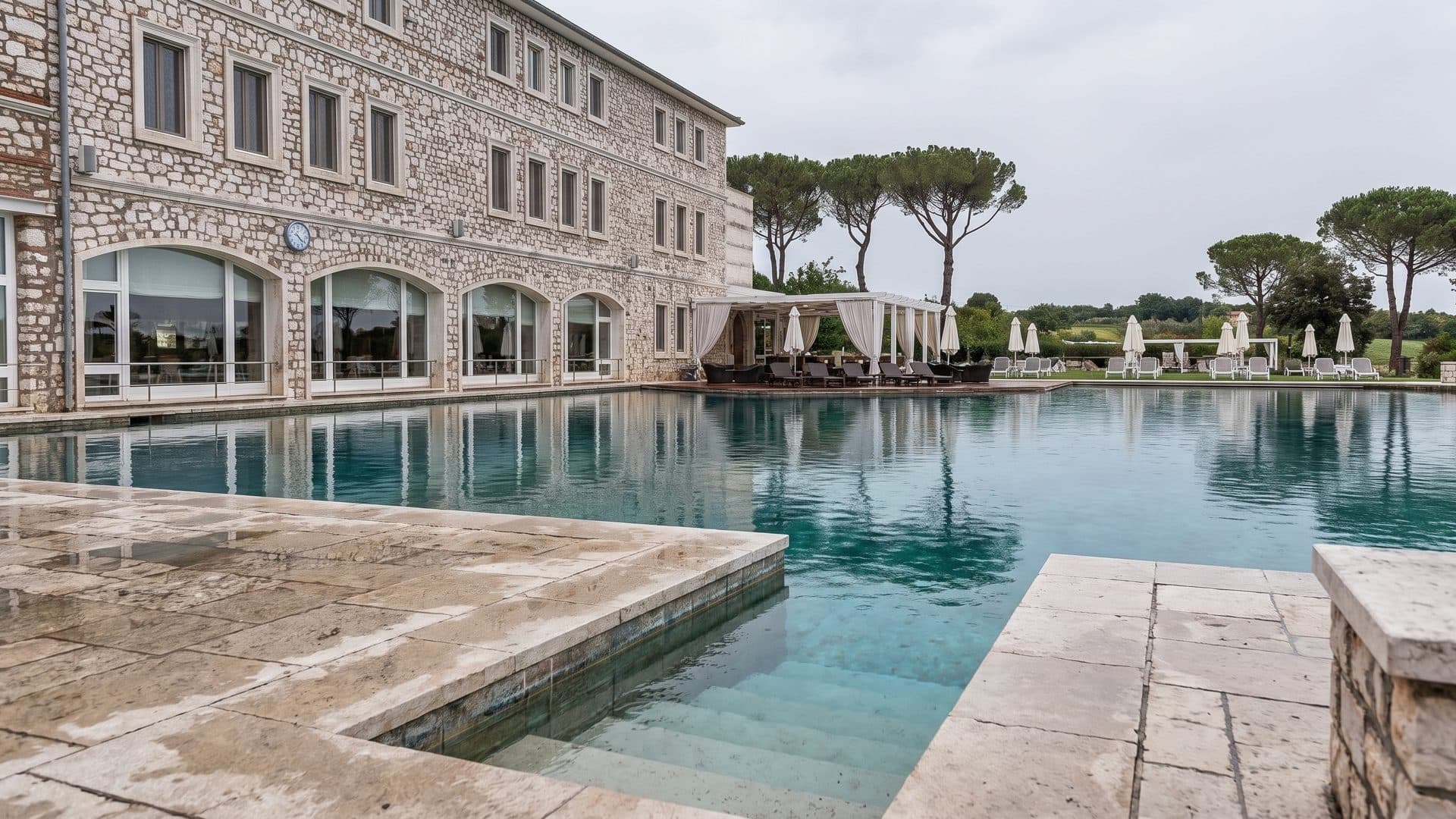 Travertine-edged thermal pool in front of the stone hotel, with umbrella pines in the distance