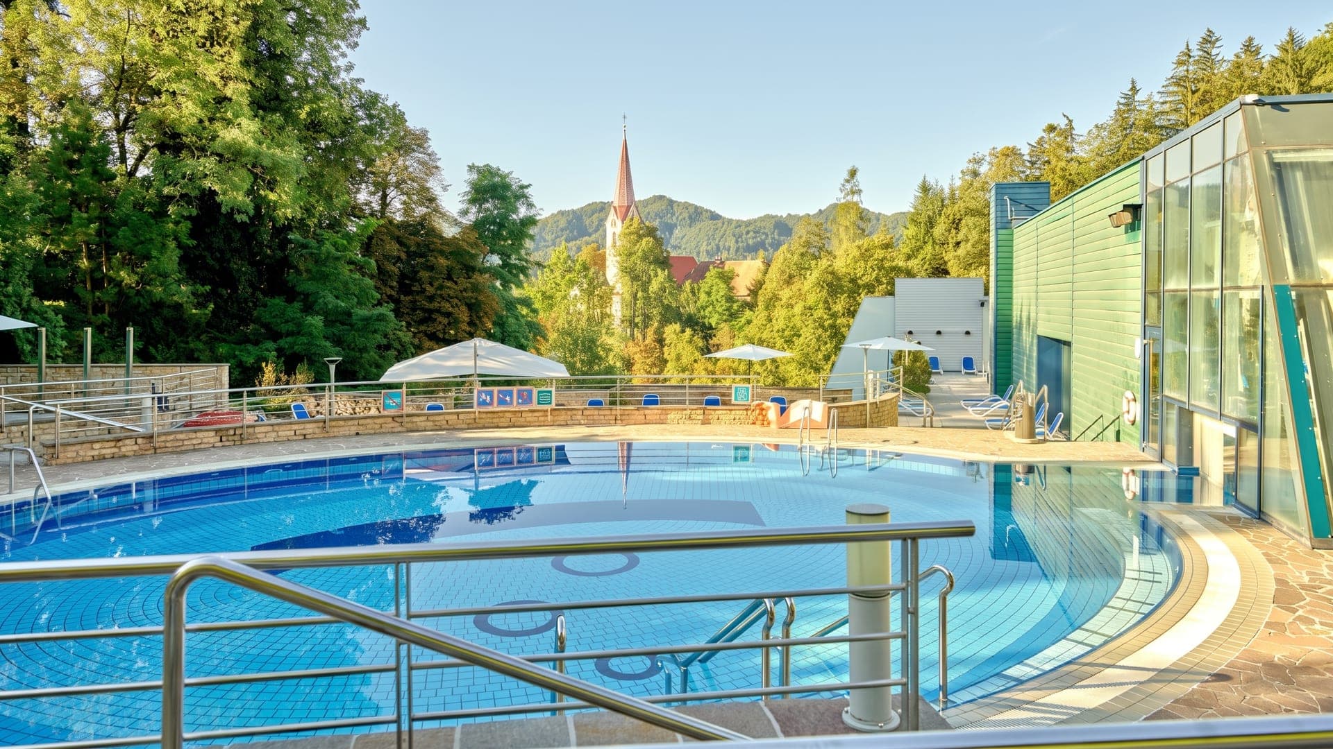 The outdoor pool at Terme Dobrna beside the glass wing of the bath house, with the village church spire and wooded hills behind.