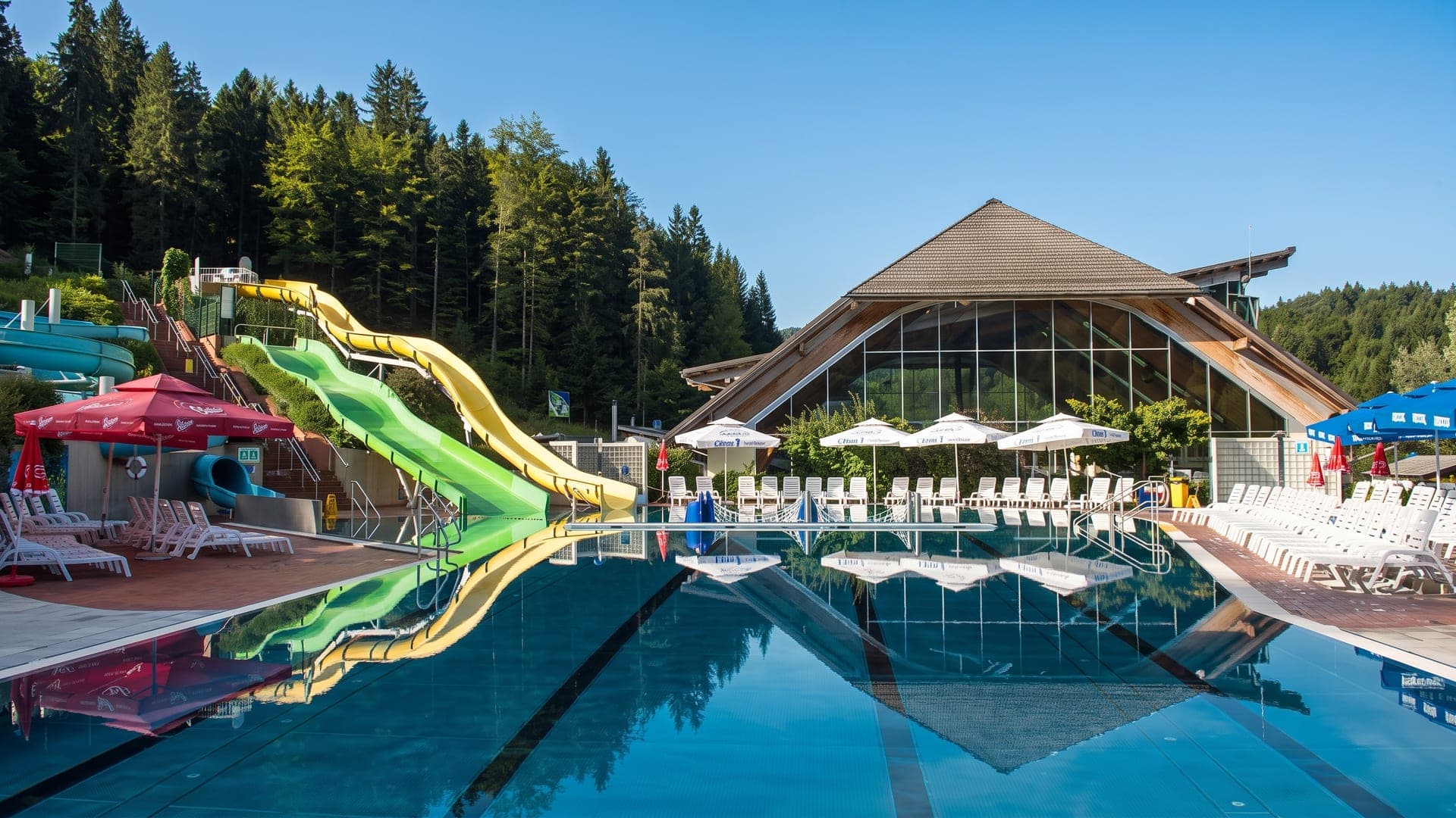 The outdoor pool at Terme Snovik, with green waterslides and the pitched timber pavilion against the pine forest.