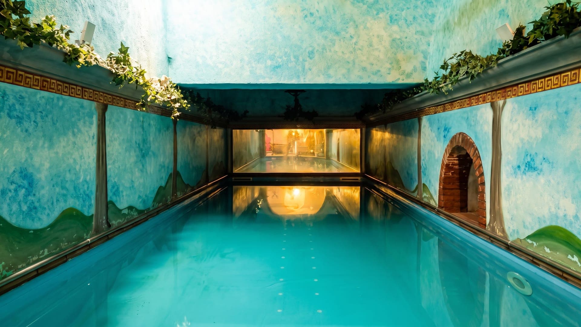 A vaulted bathing hall at the Terme Vesuviane, its turquoise pool below walls painted with a Greek-key frieze.