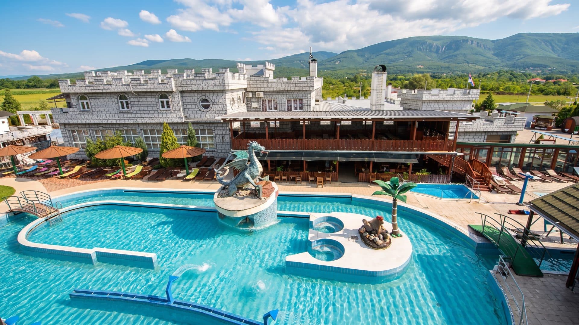 Castle-themed spa resort with a blue dragon statue rising from a turquoise pool, crenellated white walls and green mountains behind.