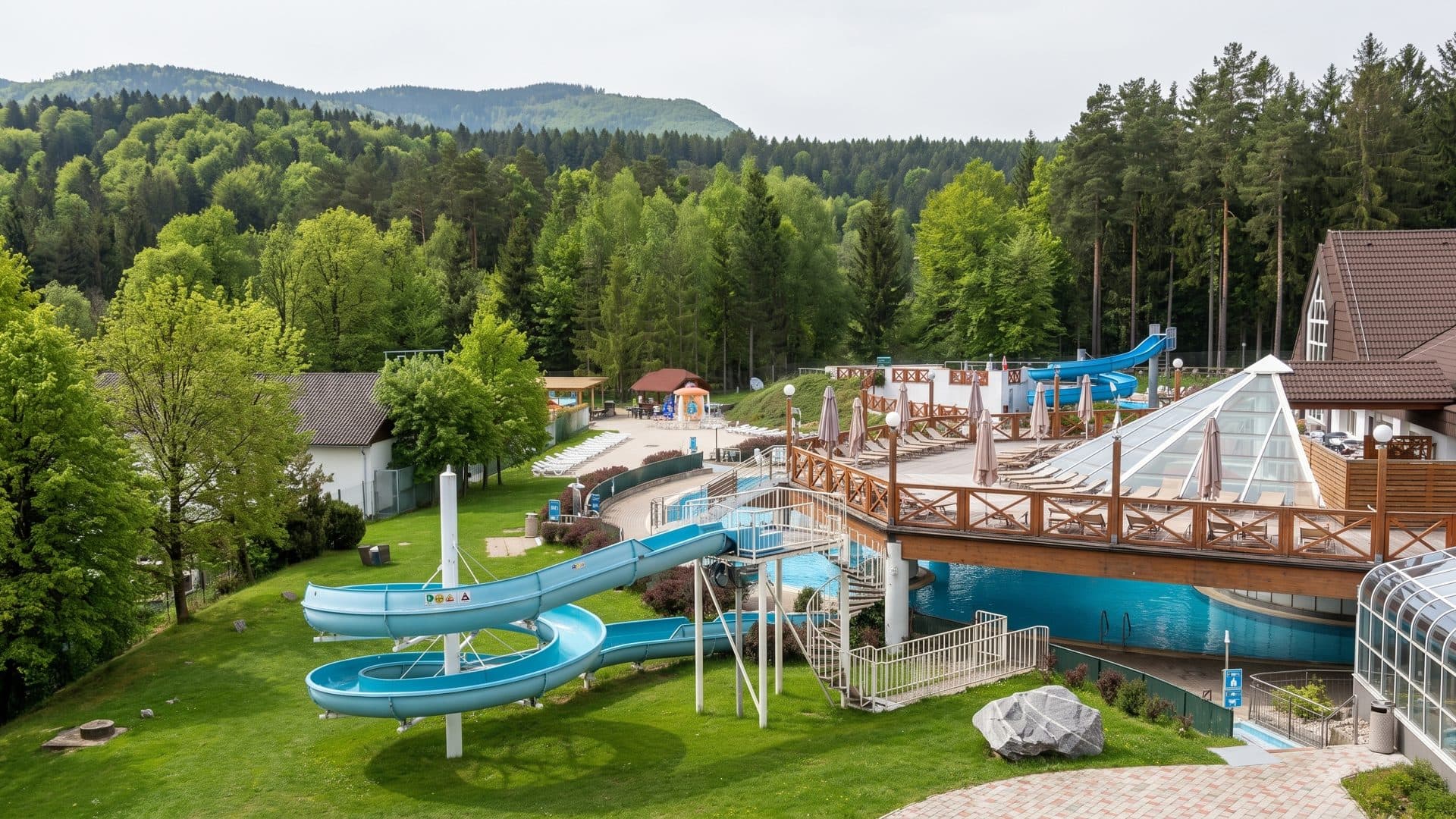 Blue water slides curving down a lawn to the outdoor pools of Terme Zrece, with timber sun decks, a glazed pavilion and wooded hills behind.