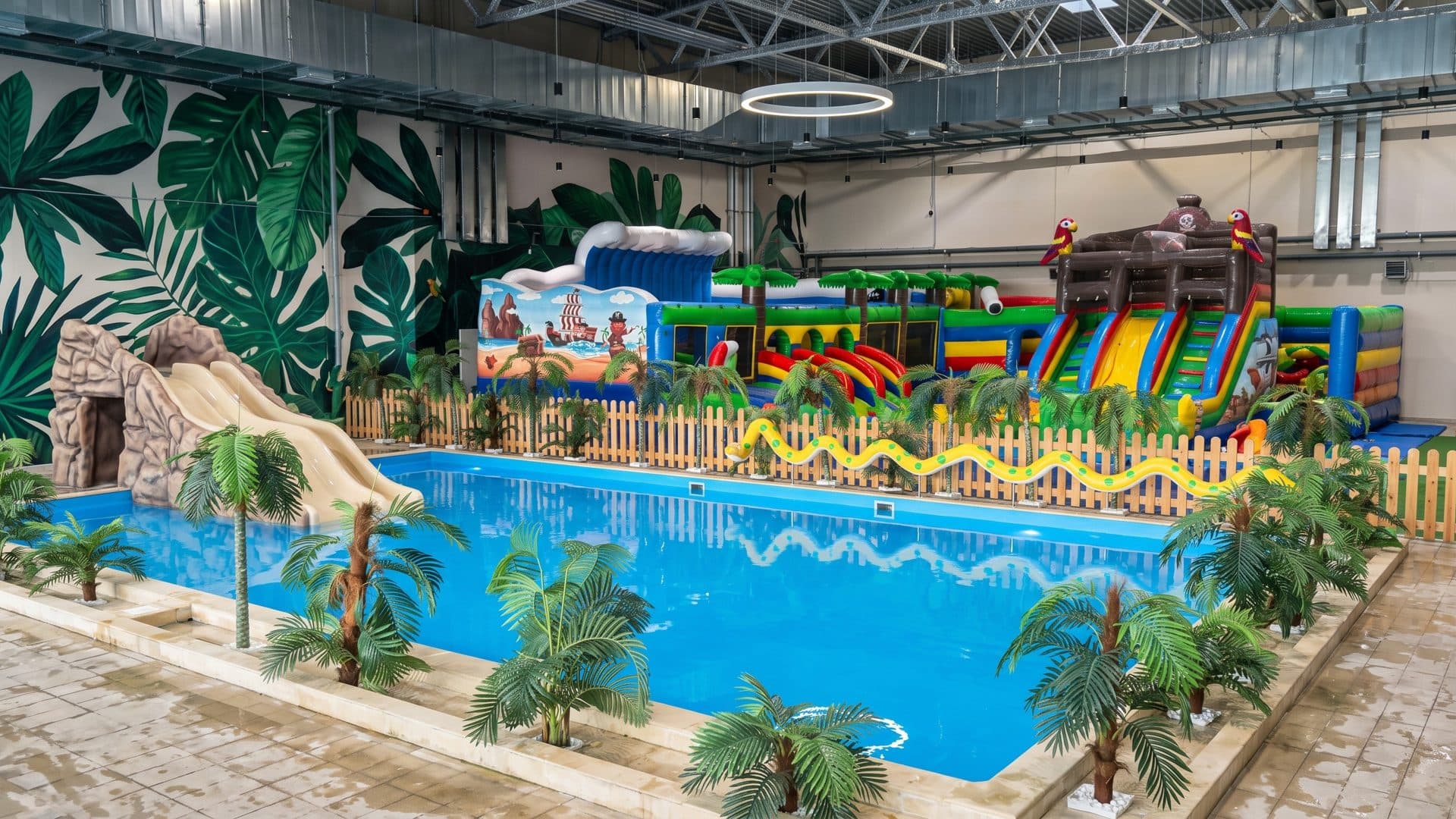 The indoor pool of Termoland with palm-leaf murals, a small slide and a bouncy castle behind a fence