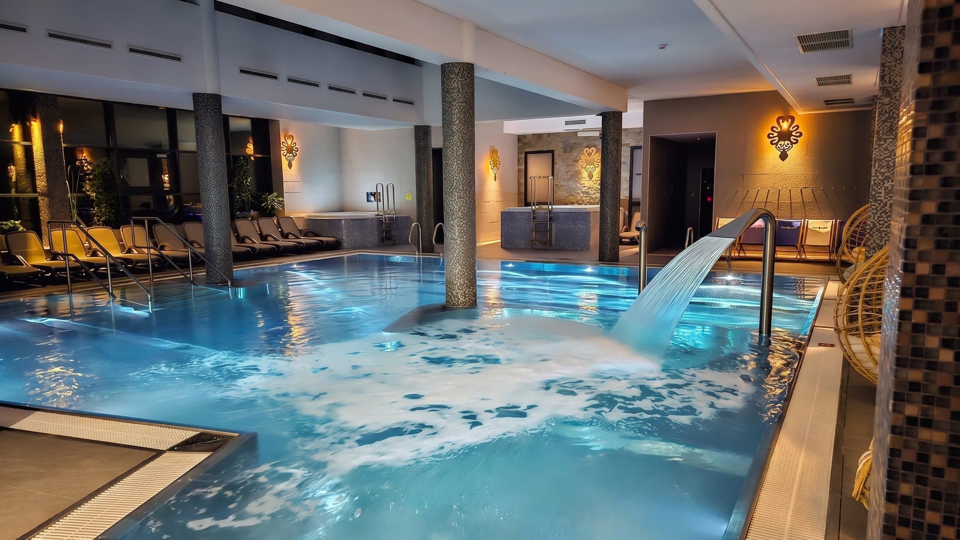 Indoor pool with a cascade jet between stone columns, warmly lit