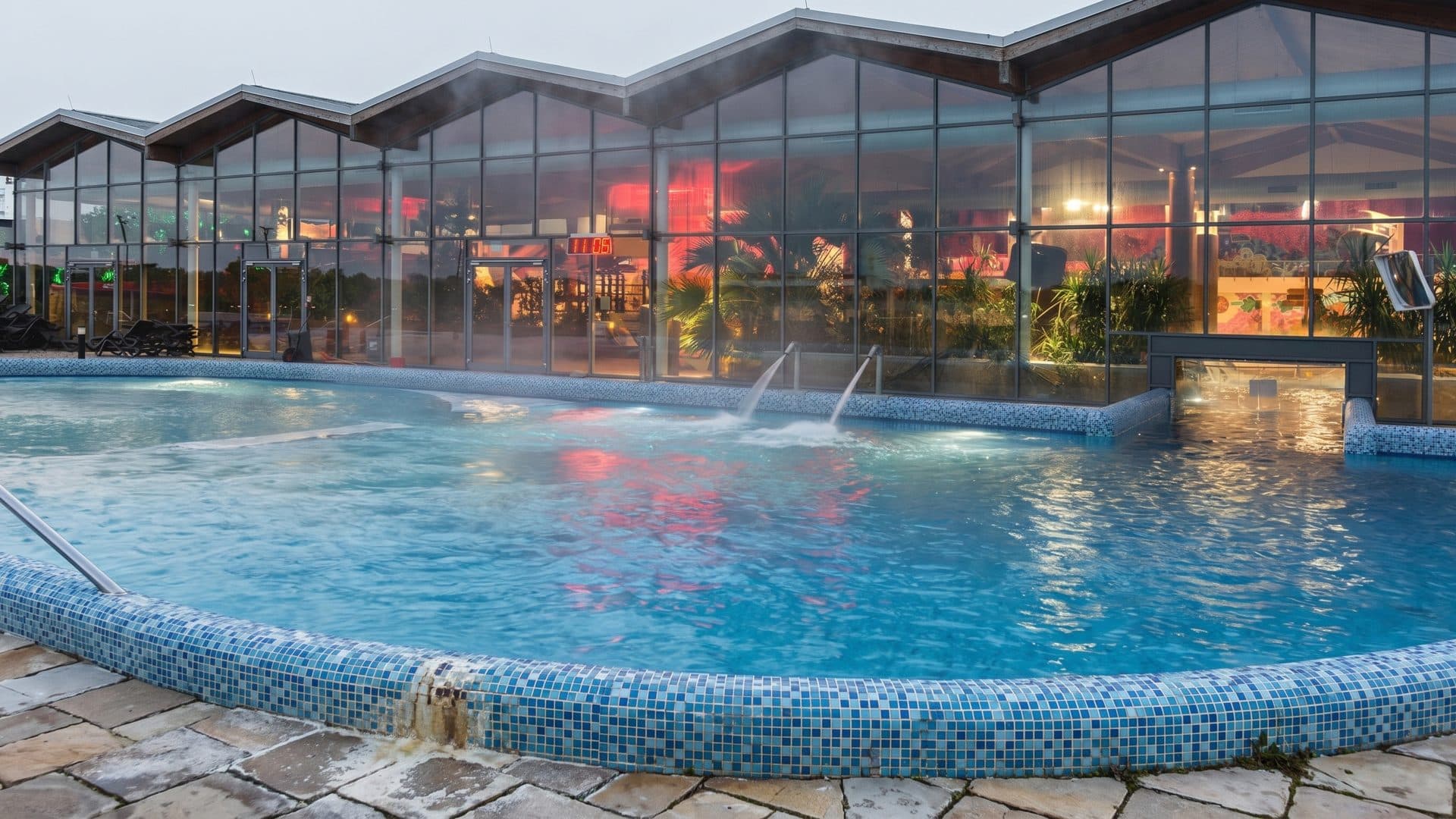 The outdoor thermal pool at Termy Maltanskie steaming in the evening, jets along its rim and the lit hall behind the glass