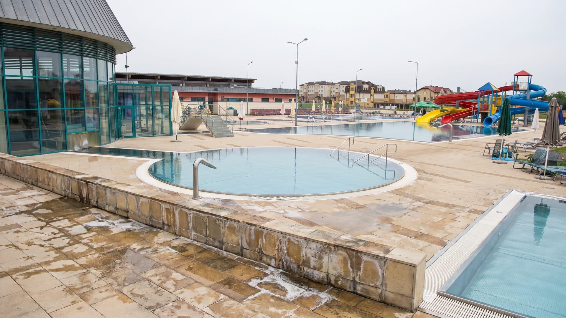 Round outdoor thermal pool on a paved terrace, with the glazed building and colourful slides behind