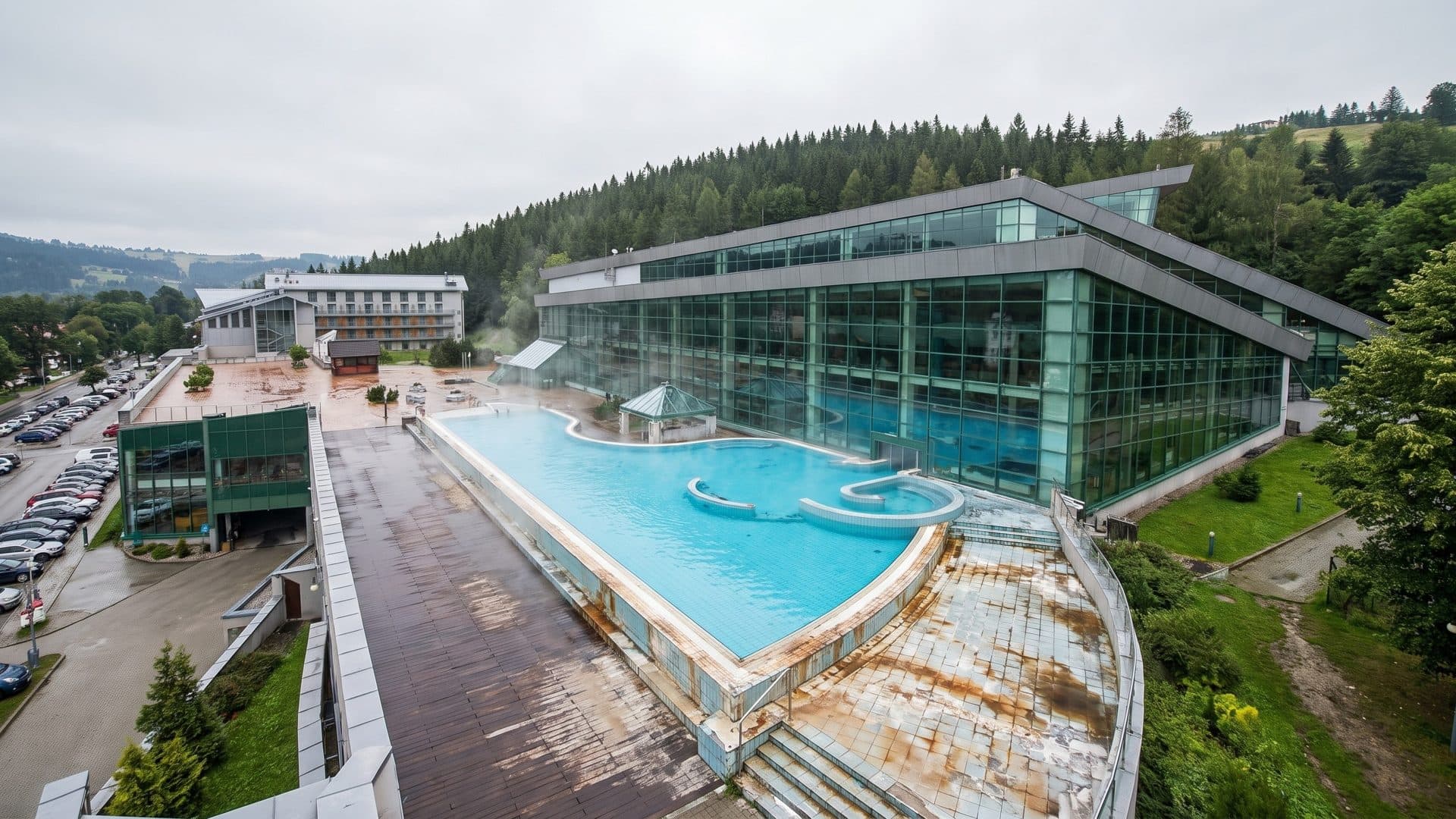 Outdoor thermal pool beside the glazed pool hall, seen from above with forested hills behind