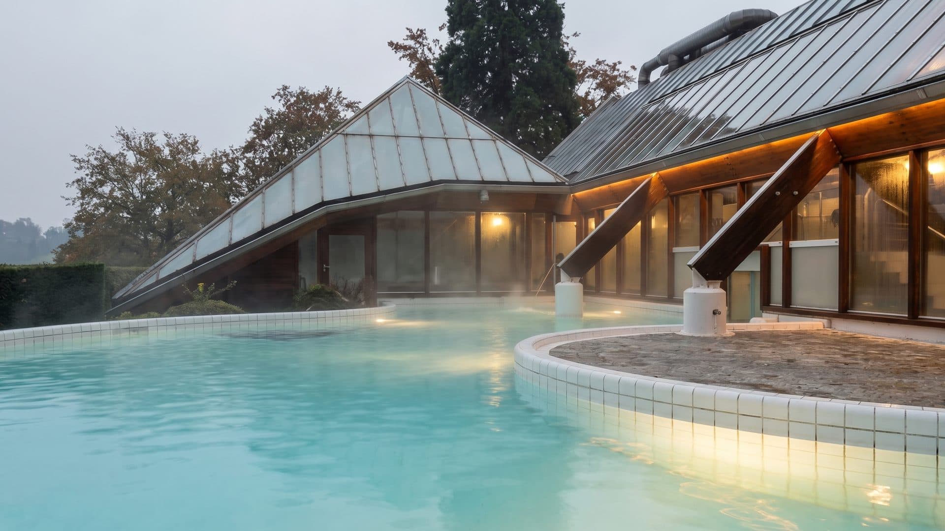 The outdoor thermal pool at Thermae 2000 steaming at dusk, the curved building's lit windows above it
