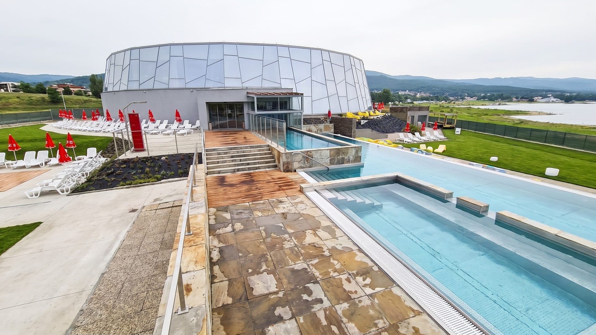 Outdoor pools and a stepped terrace leading to the round glass rotunda, with red parasols and the lake
