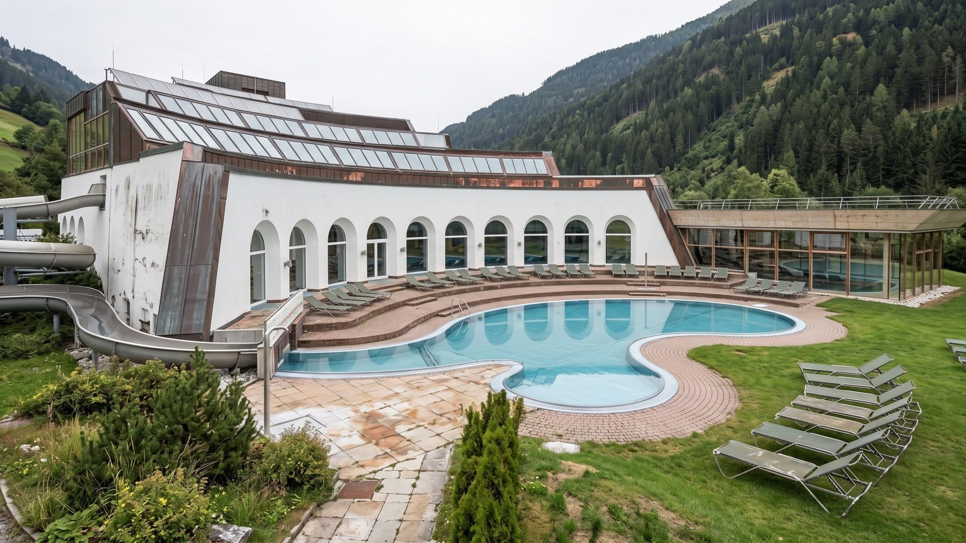 The curved outdoor pool of the Thermal-Romerbad in front of the white arcaded bath hall, forested mountains behind