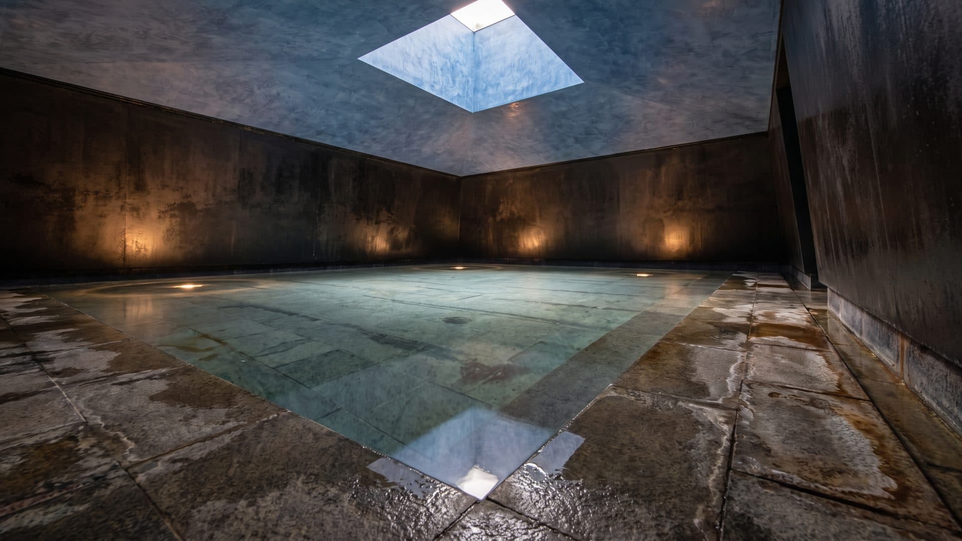 A vaulted concrete bathing room at Thermalbad Zurich, the shallow pool lit from below and a skylight overhead