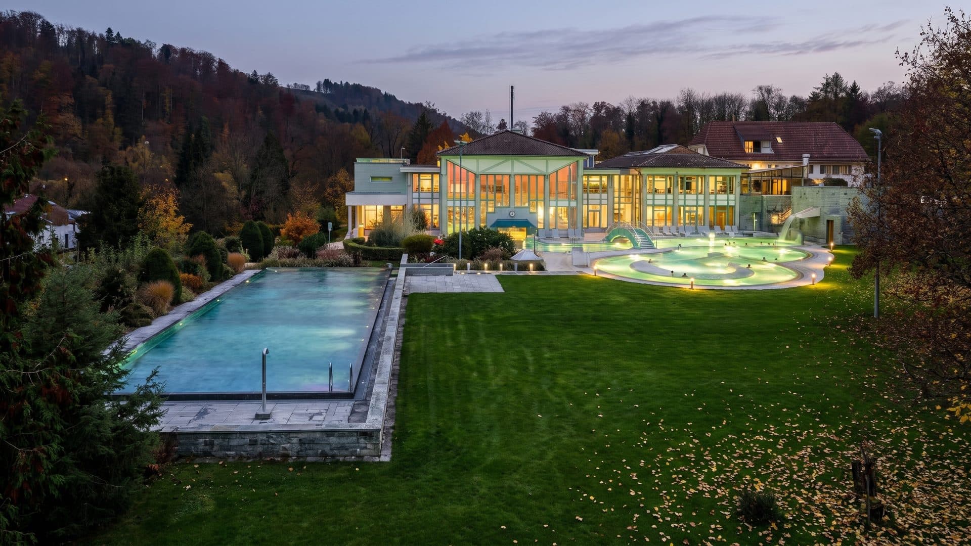 Thermalbad Schinznach at twilight: the outdoor pools on their terraces in the wooded park, the bath buildings lit along one side.