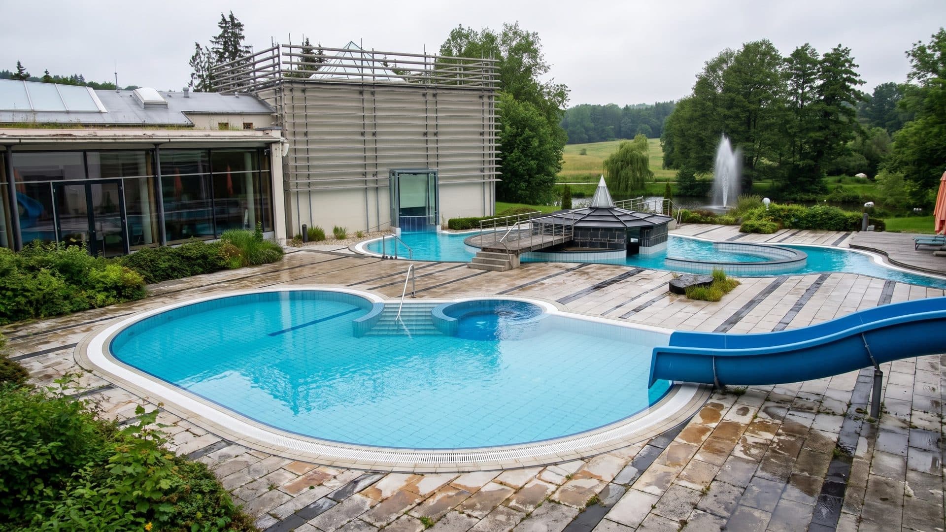 Outdoor thermal pool with a blue slide, a pavilion island and a fountain in the pond beyond