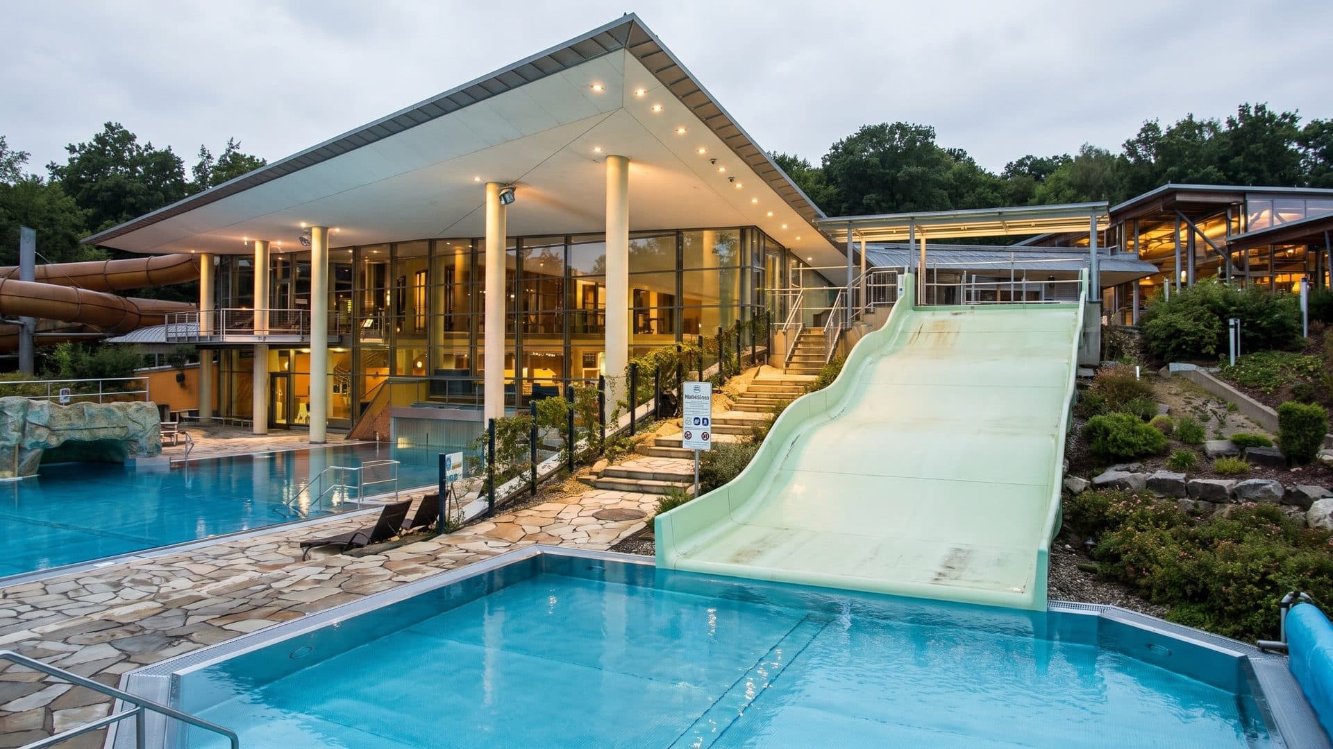 Outdoor pool with a pale green wide slide running down beside the lit timber-roofed pavilion