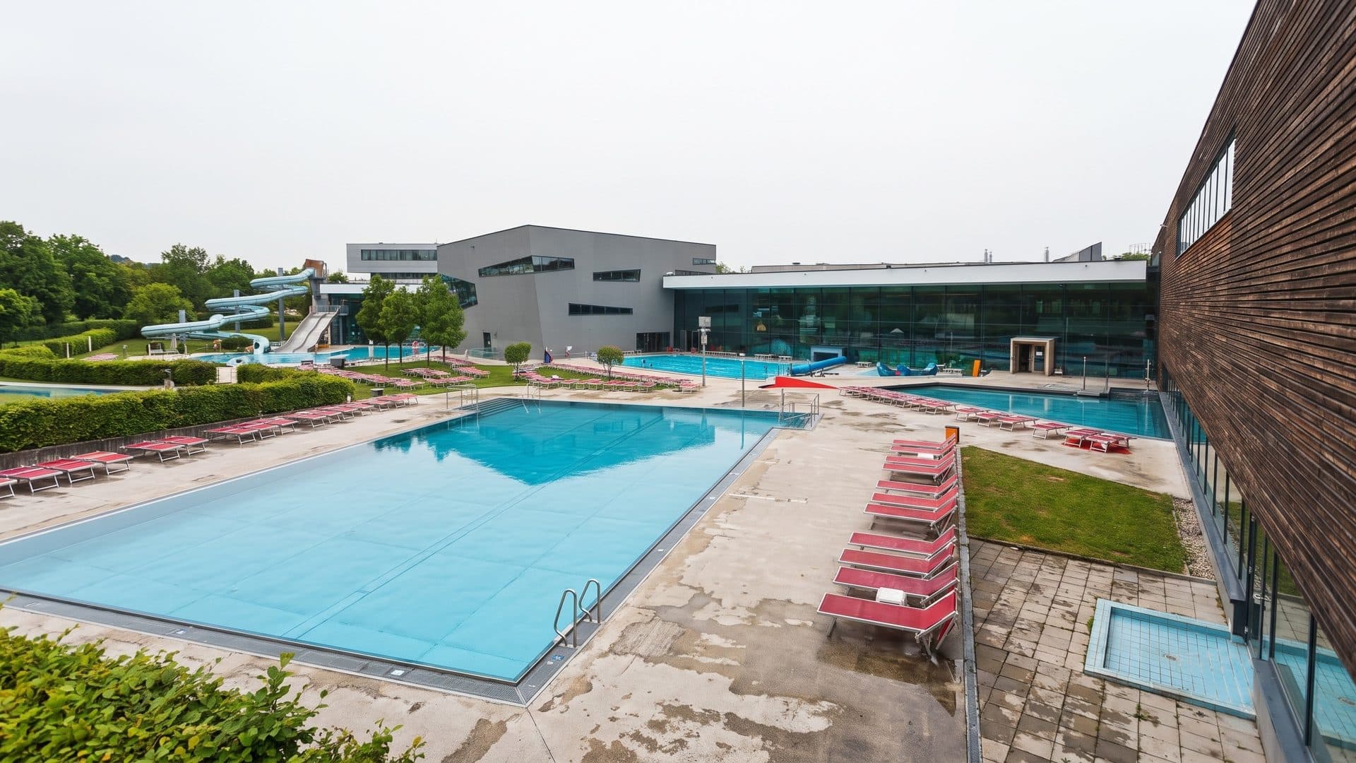 Outdoor lap pool lined with red loungers between the timber-clad building and the grey halls
