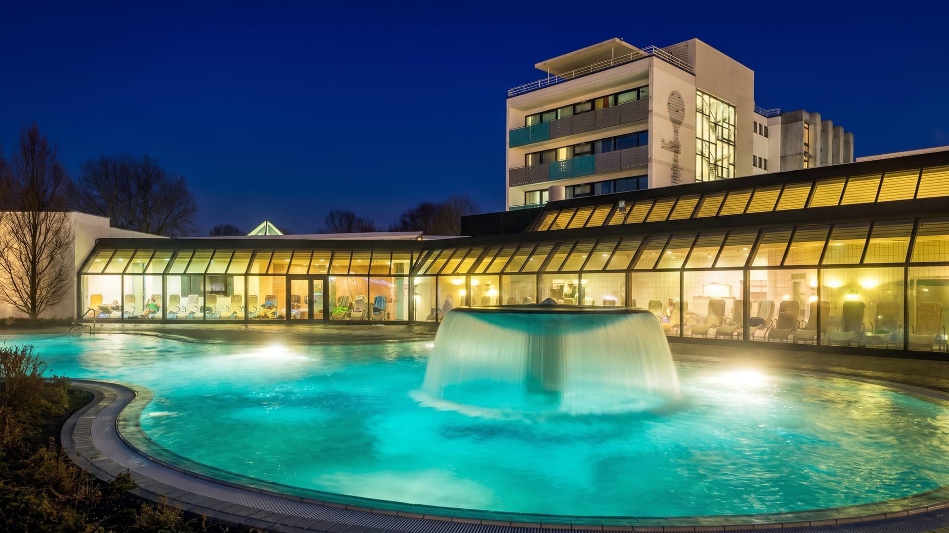 The outdoor pool at the Adelindis Therme in Bad Buchau at night, a large mushroom fountain pouring into the turquoise water.
