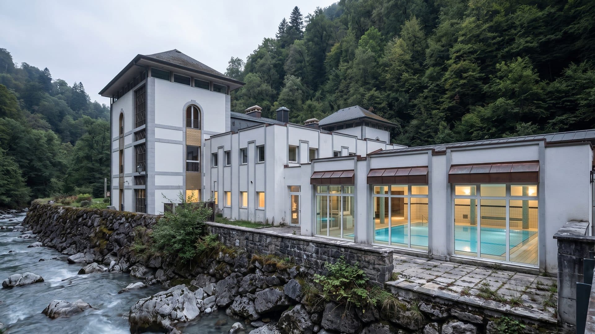 The white bath building of Saint-Gervais-les-Bains standing over its gorge, the lit indoor pool visible through the glazed front and wooded slopes above