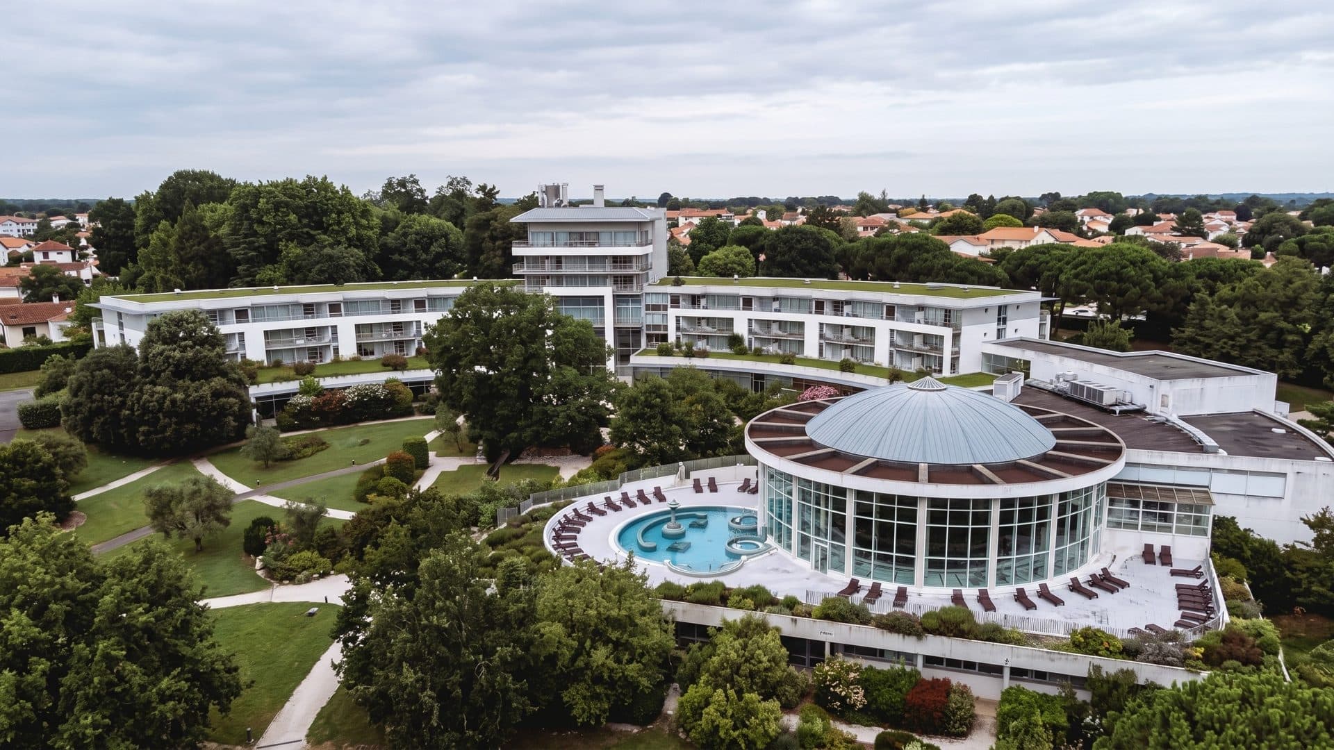 Aerial view of the Sourceo thermal baths: a glazed rotunda with a round outdoor pool, set in a park below the white hotel wings