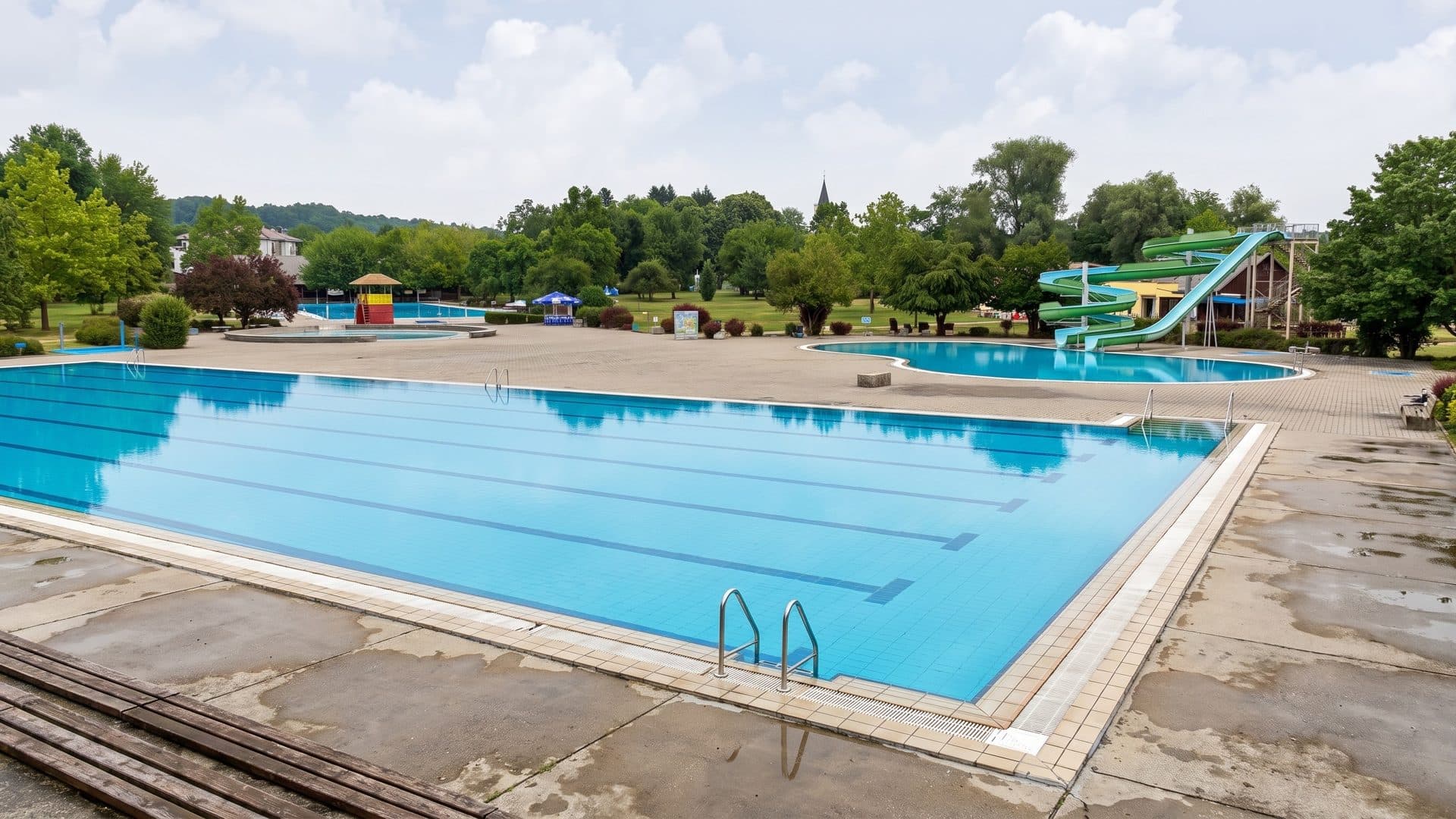 Large outdoor pool on a wide deck in a park, with a green slide and a children's pool beyond