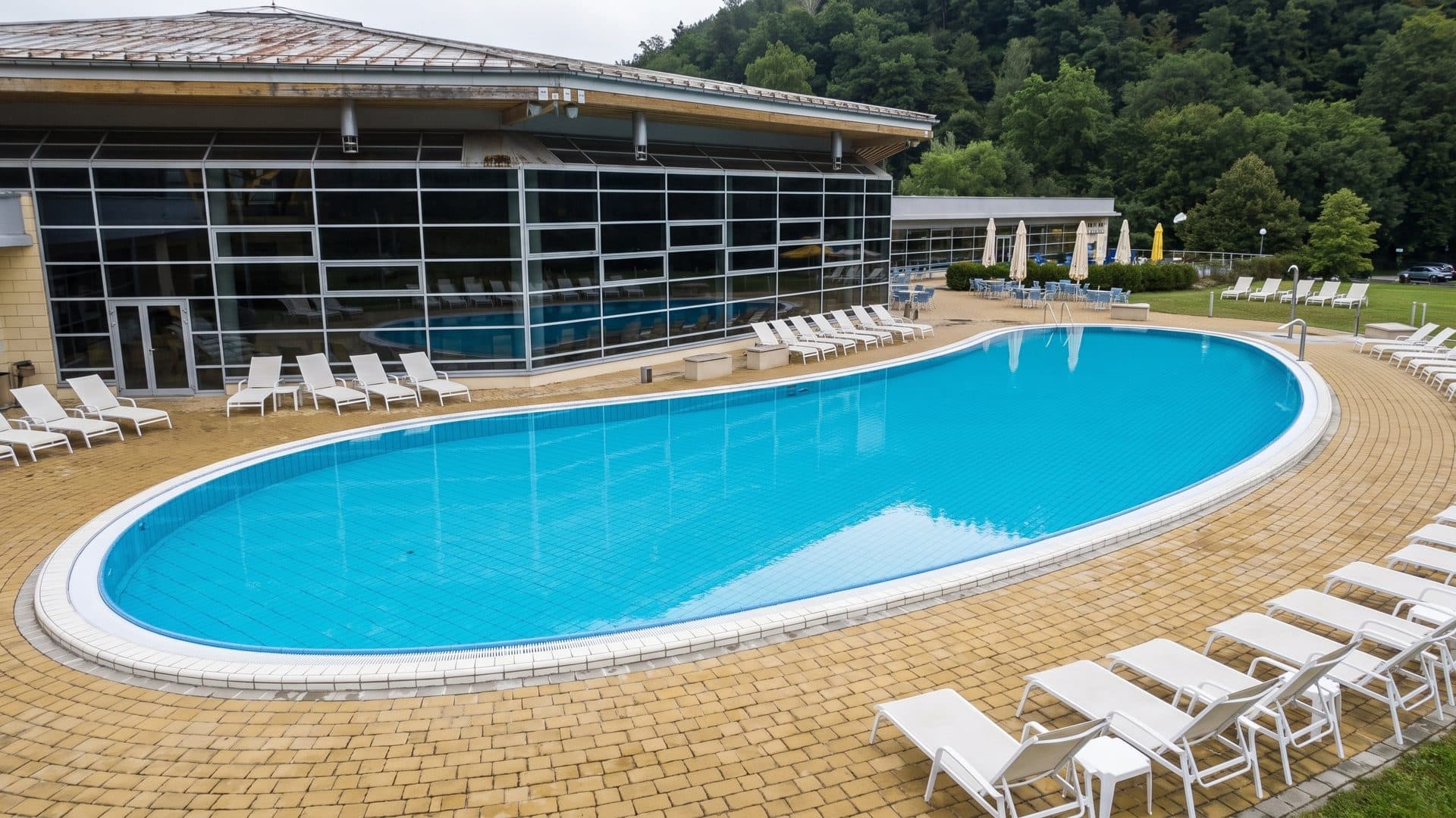 Kidney-shaped outdoor pool with white loungers, beside the glazed hall below wooded sandstone hills