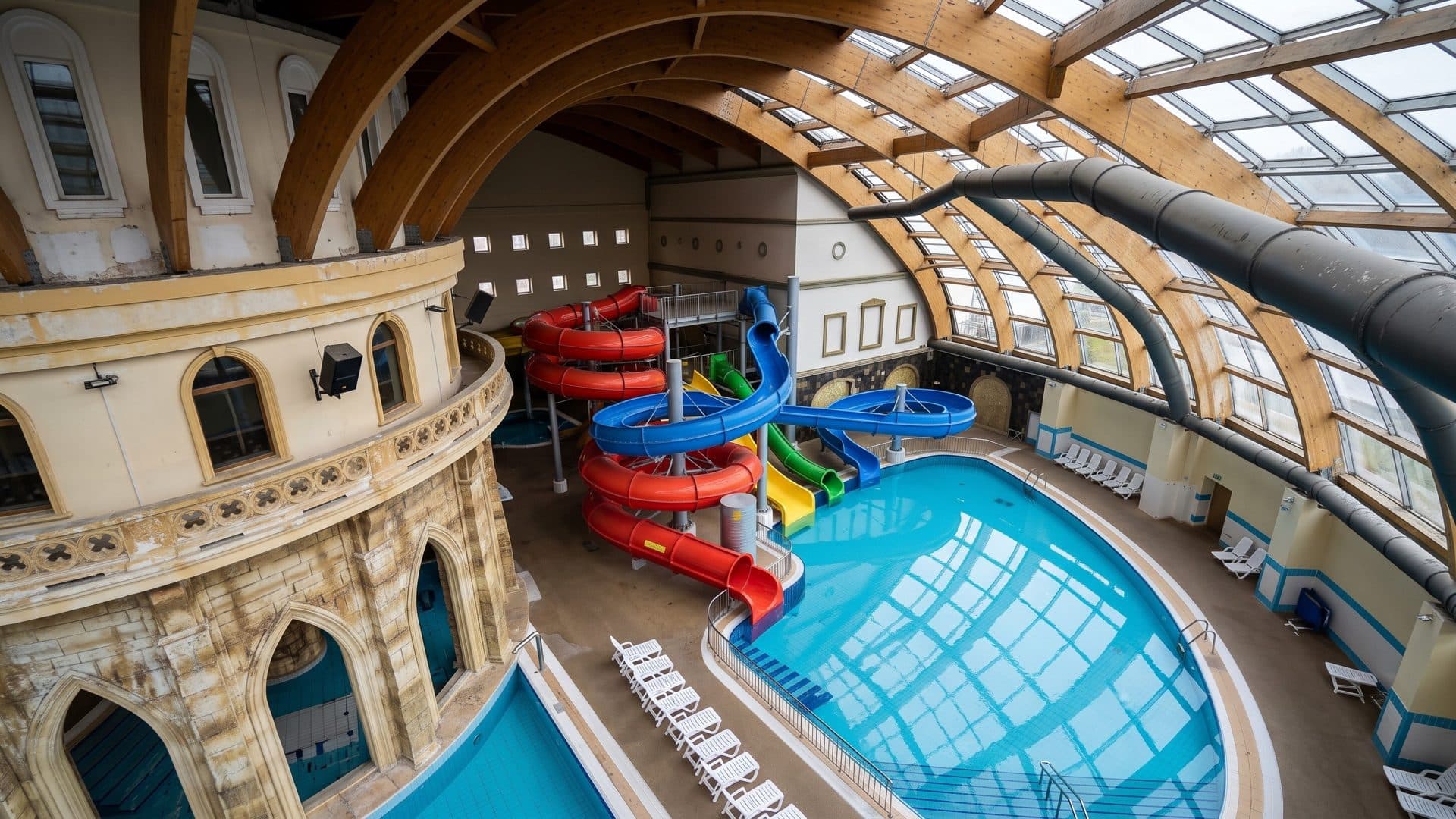 Red and blue slides over the pool under a glazed barrel roof at the Karibiya family centre, a colonnaded facade alongside