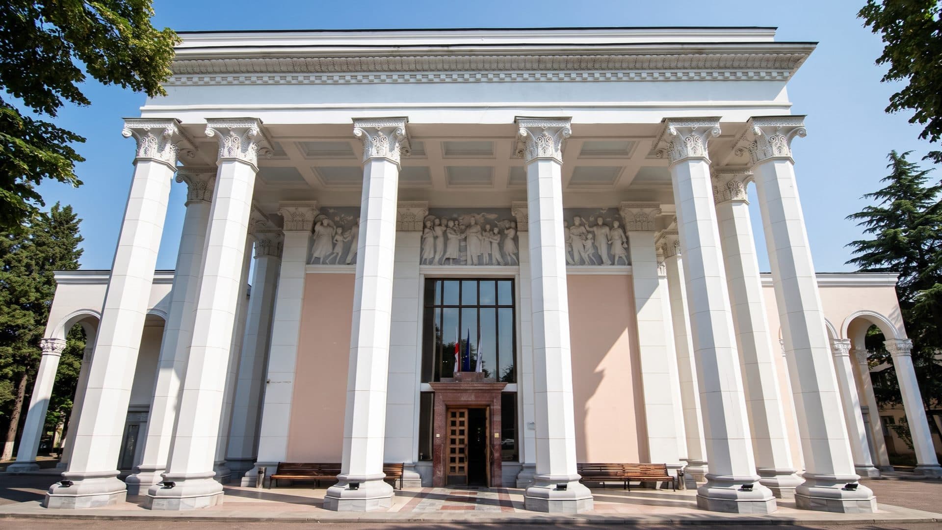 Grand white neoclassical bathhouse with tall Corinthian columns and a sculpted frieze, set among trees at a Georgian balneological resort.