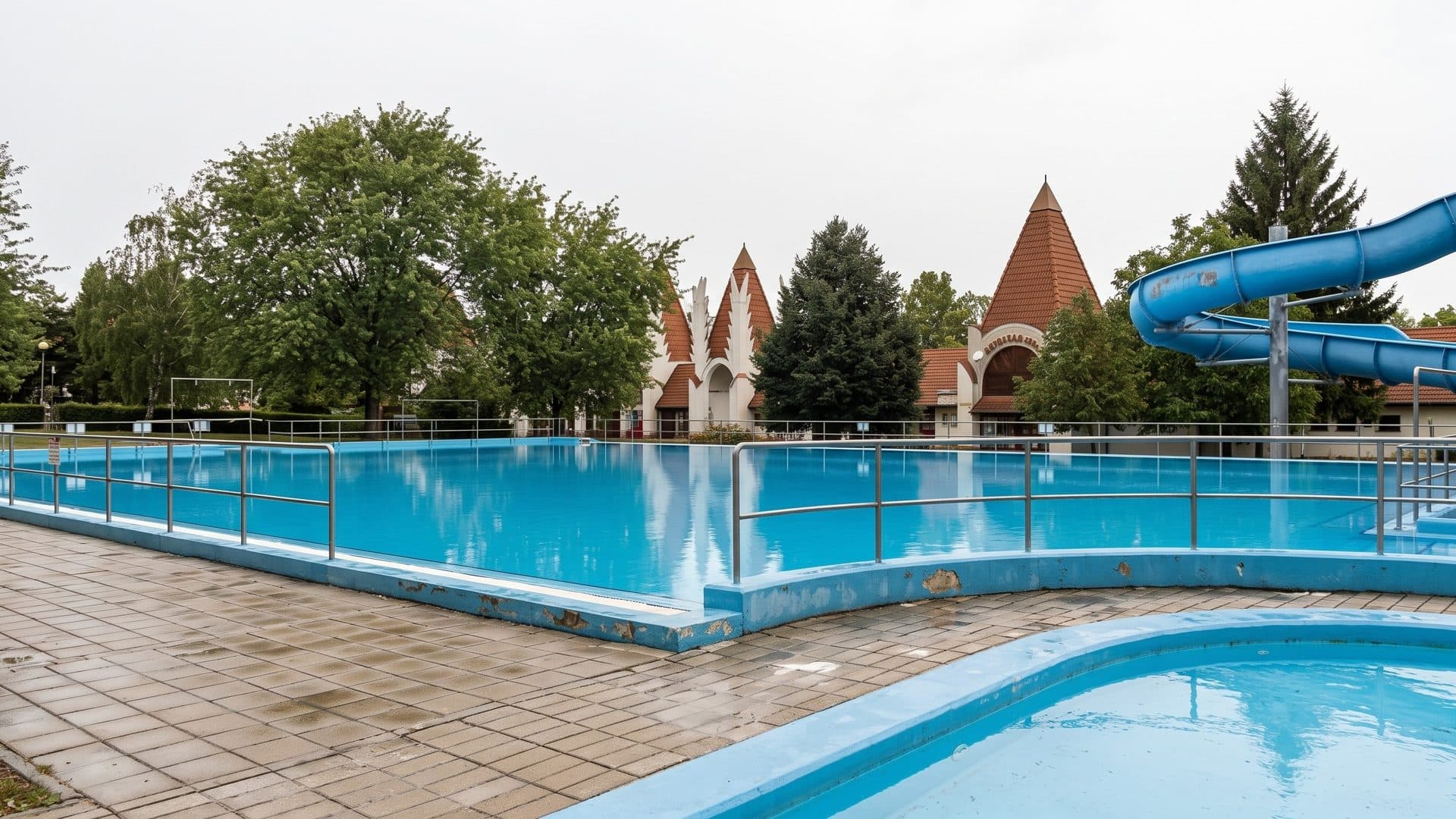 The large open-air pool of the Vegardo baths with a blue tube slide on the right and pointed turret roofs behind the trees