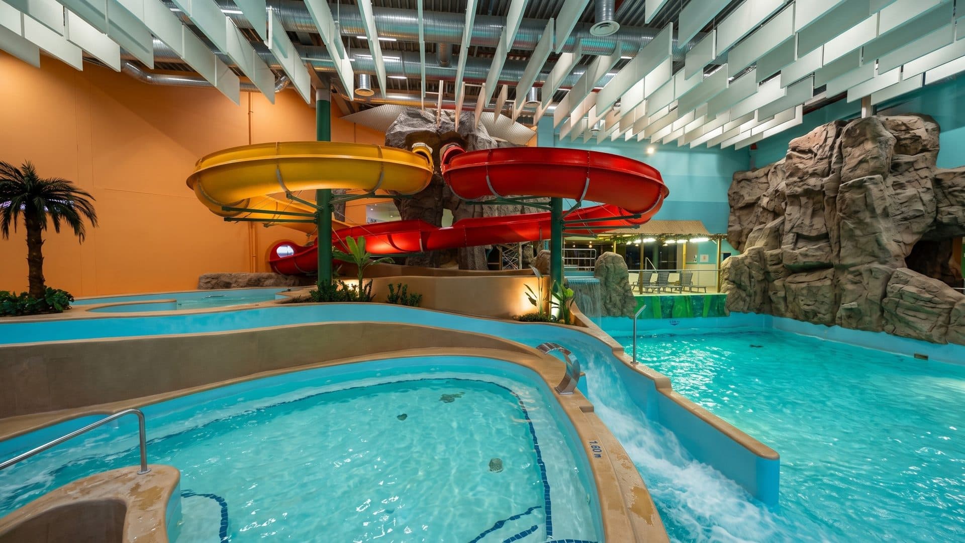 Yellow and red slides curving over the indoor leisure pool of the Ventspils water park, an artificial rock beside it
