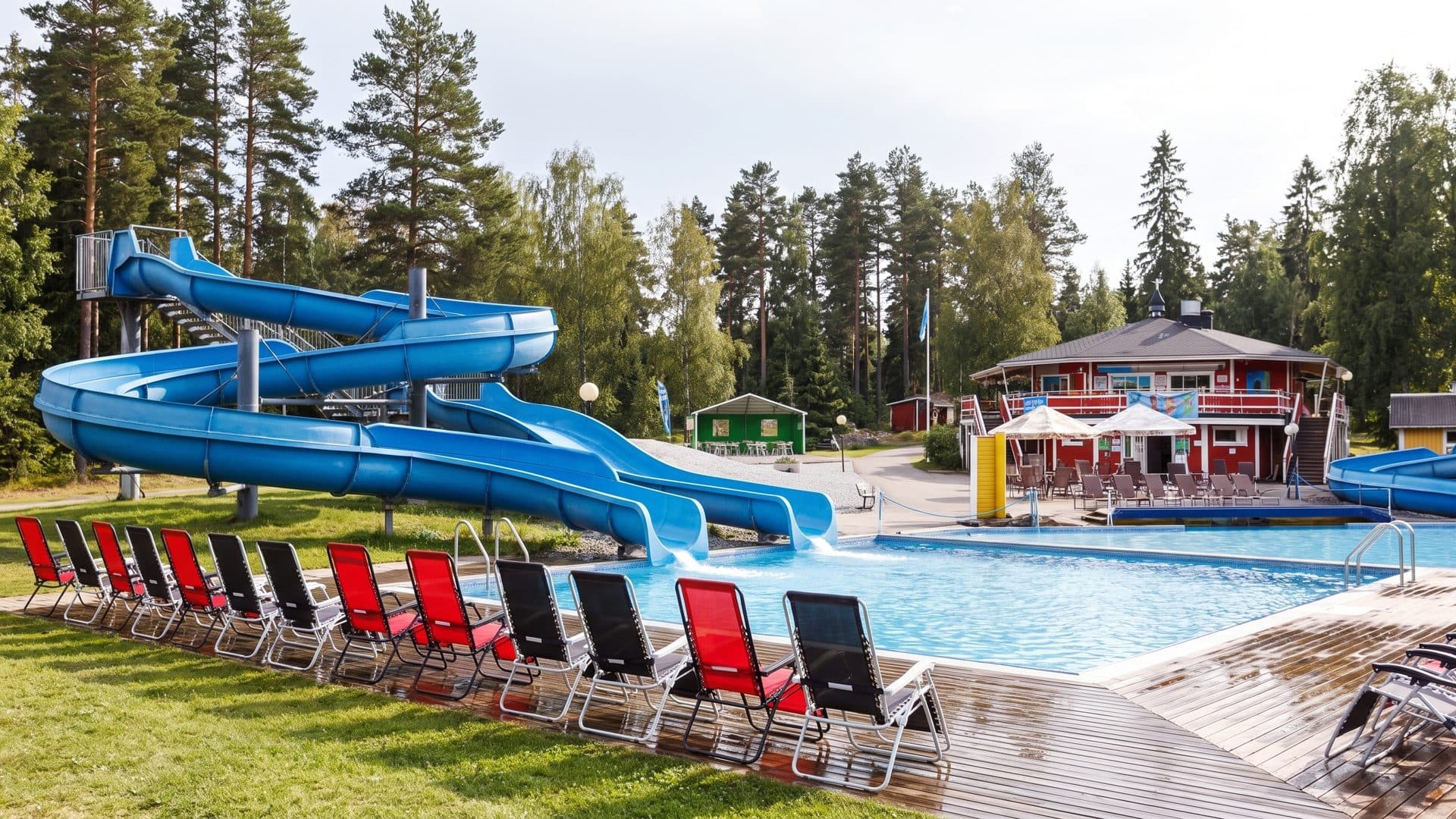 The blue water slides at Kesamaa standing against the pines, with red loungers along the near edge of the outdoor pool and the red clubhouse across the water