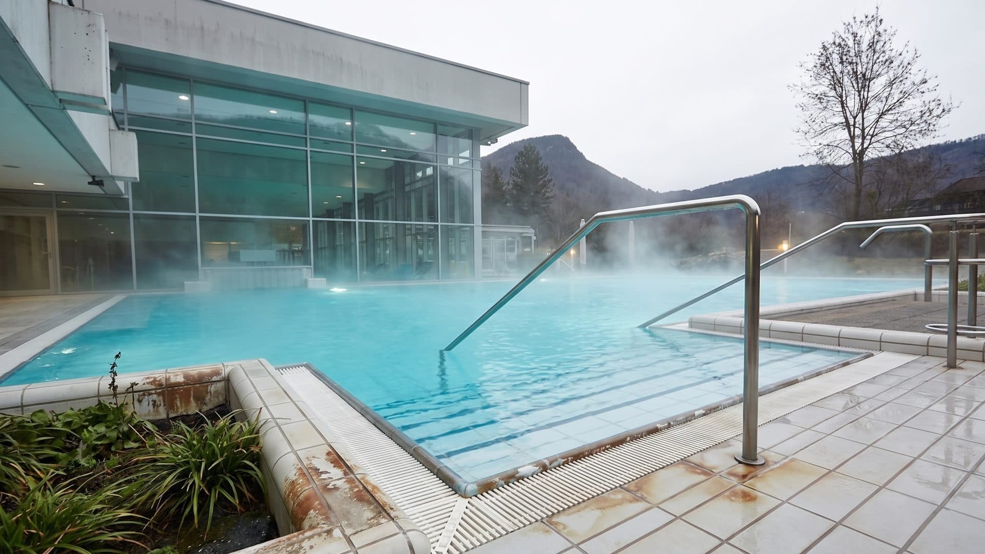 Steaming outdoor thermal pool with steel handrails, the glazed bath house and mountains behind
