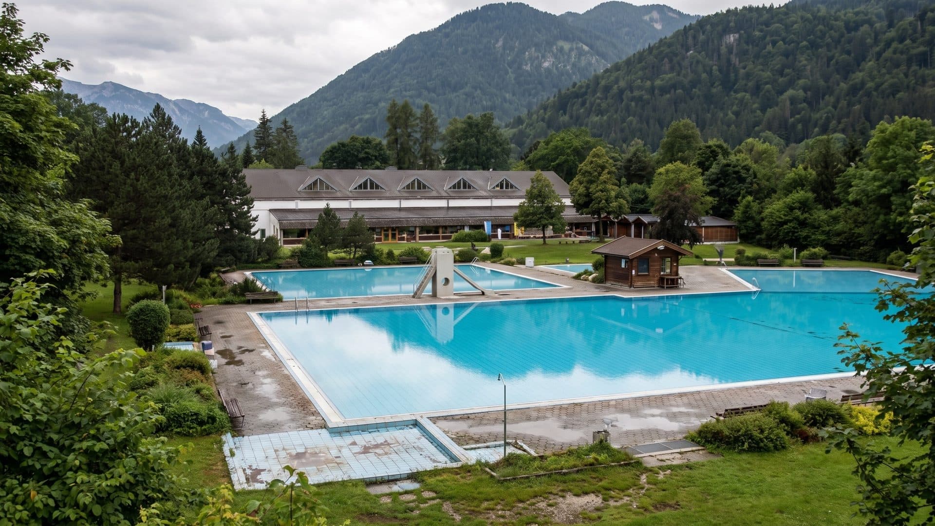 Outdoor swimming pool in a green valley, with the long bath building and wooded Alpine peaks behind