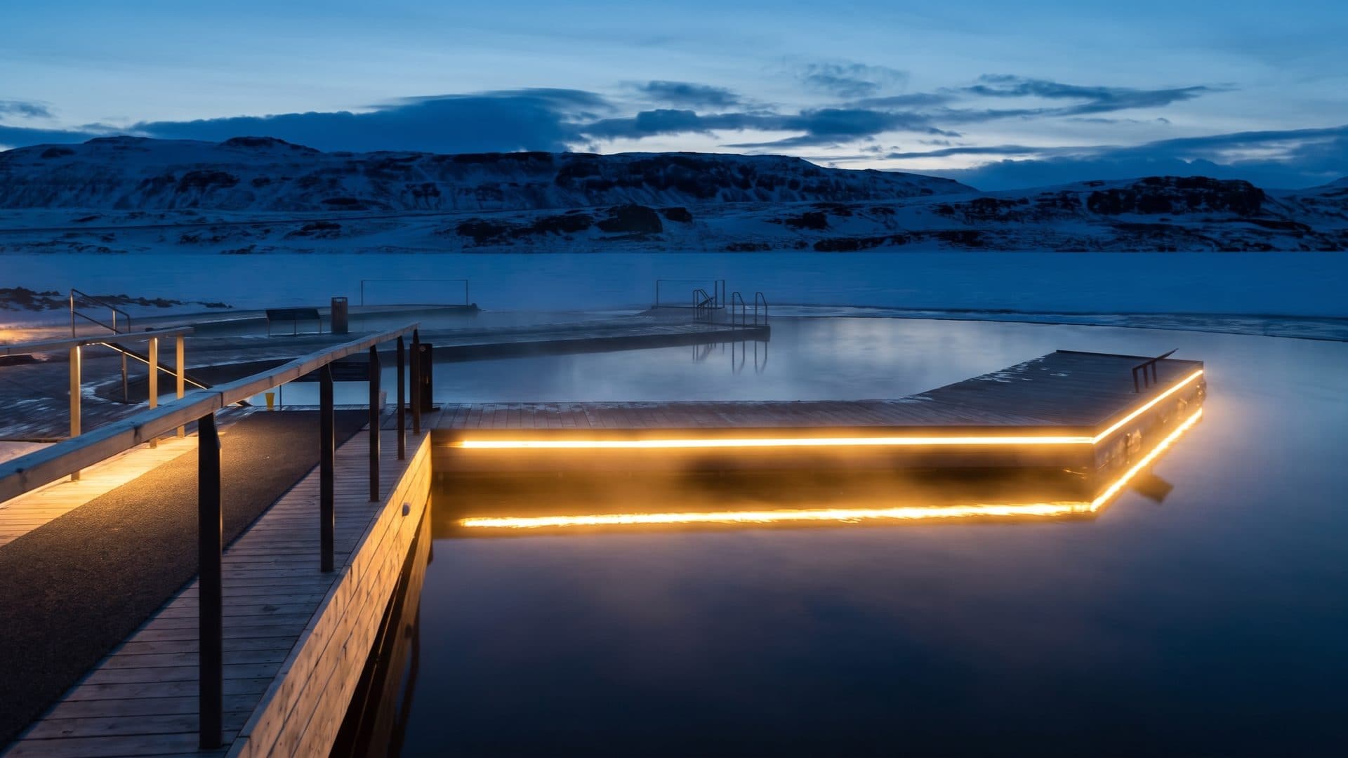 The floating lake pools at dusk in winter, warm strip-lighting glowing along the decks against snow-covered mountains.