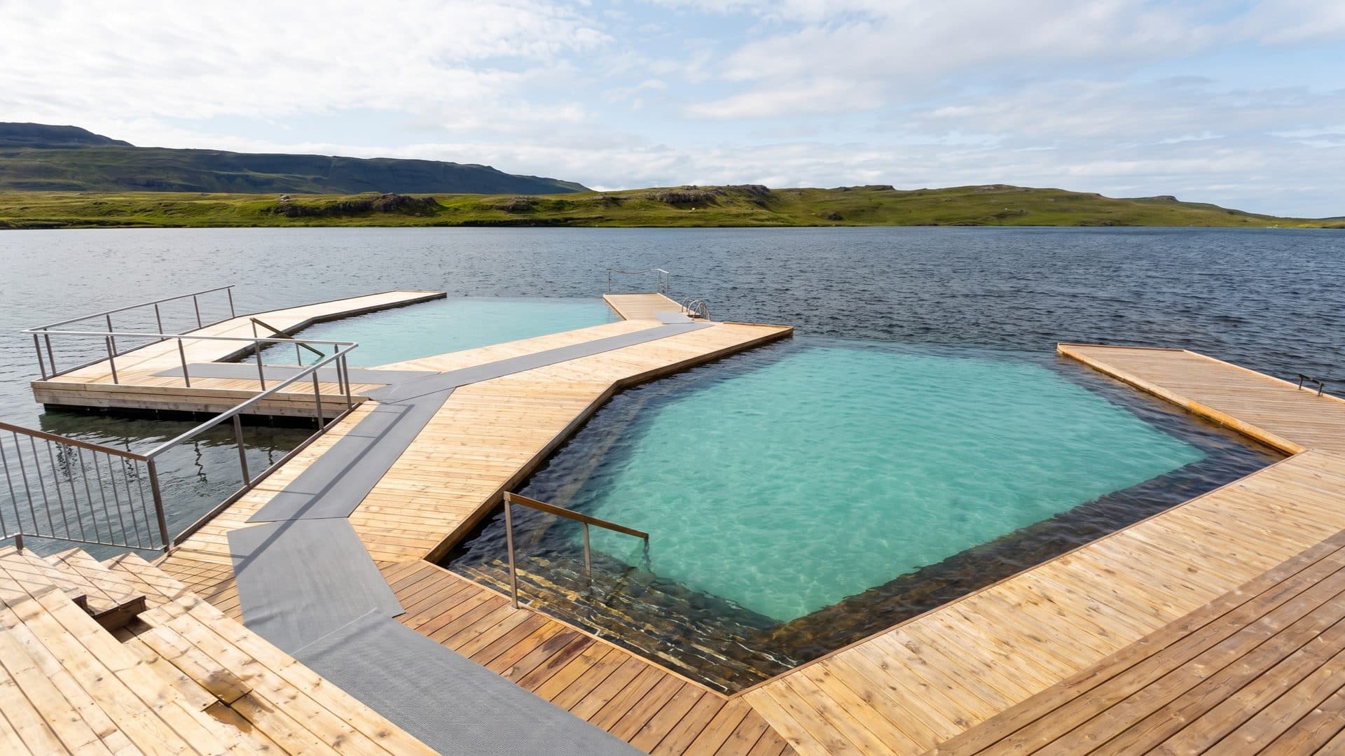 Floating geothermal infinity pools set into a cold lake with timber decking, teal water against green treeless Icelandic hills.