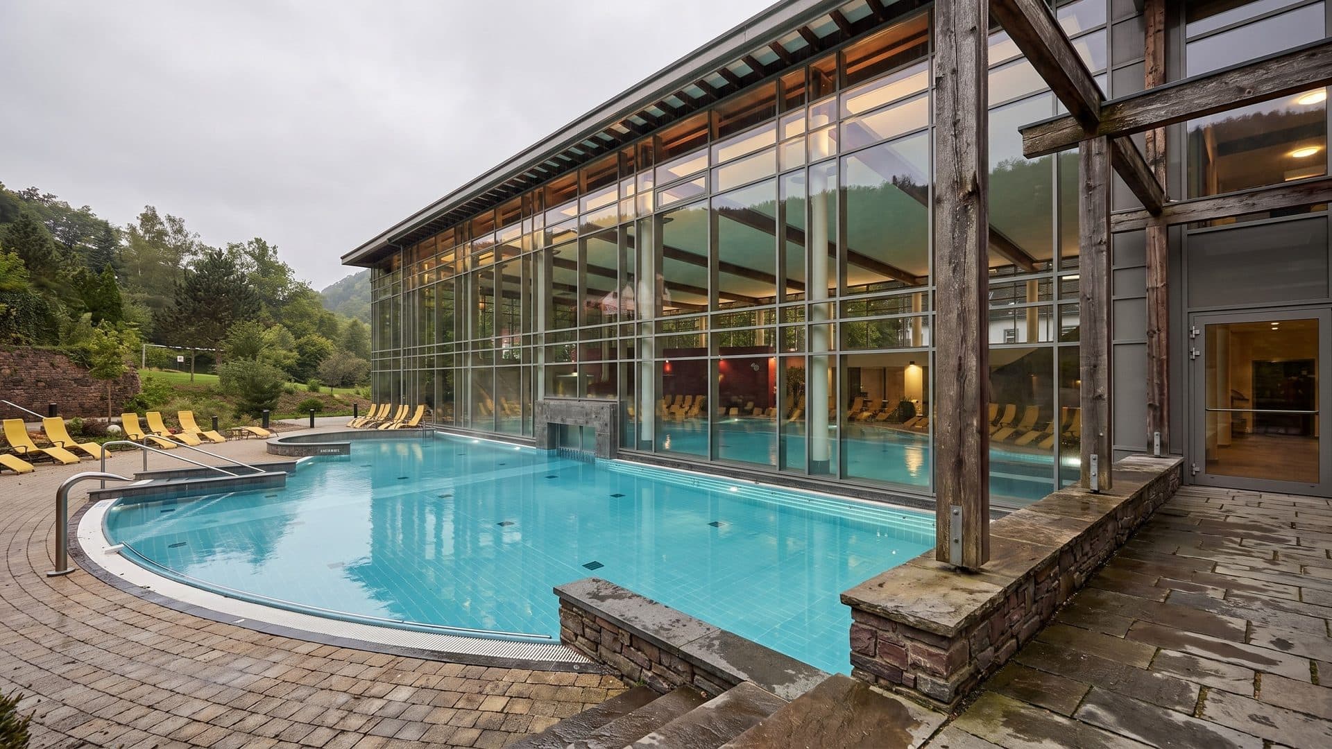 Outdoor thermal pool curving around the timber-and-glass bath house, with loungers on the terrace