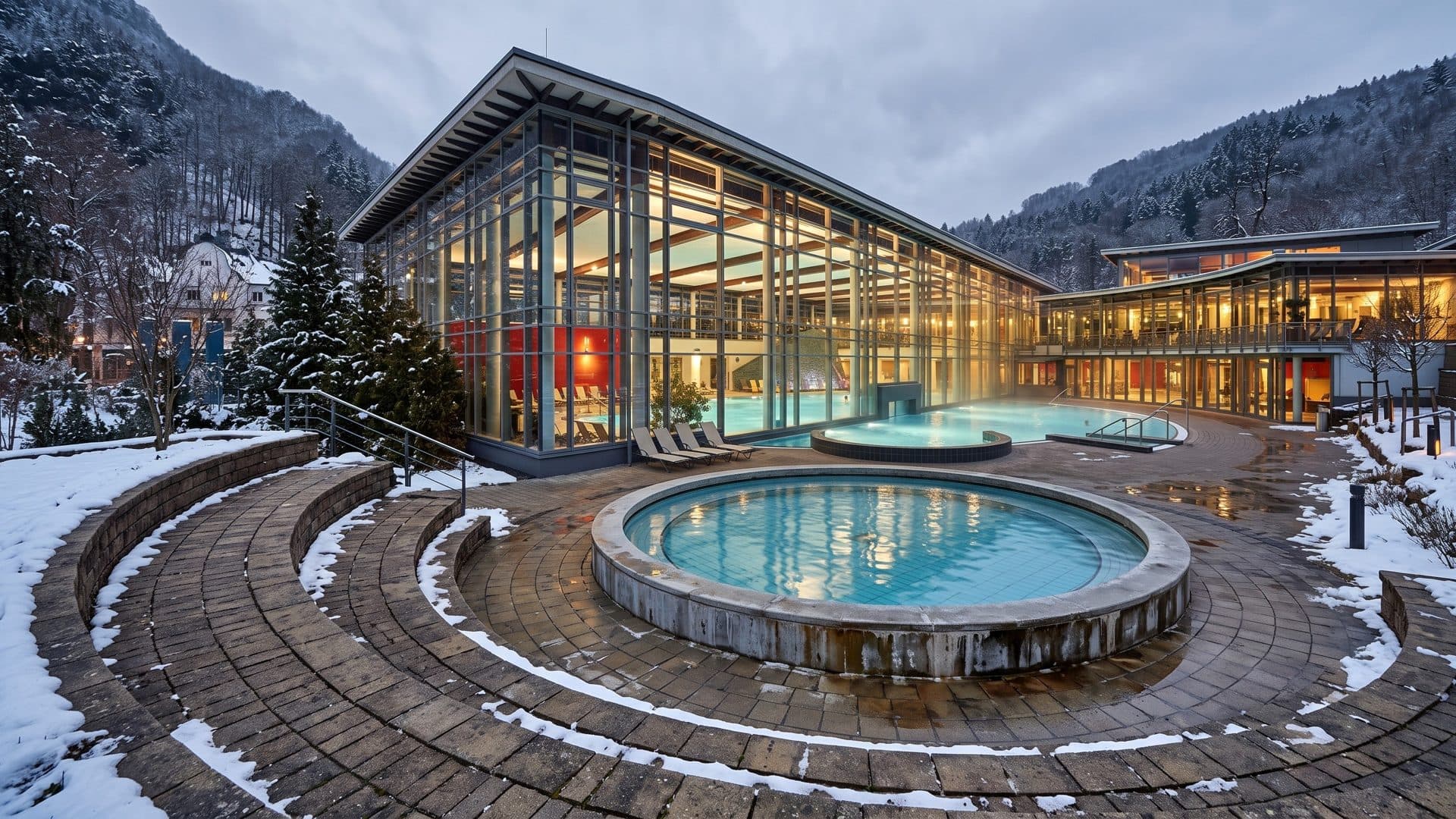 The round outdoor pool of the Vulkantherme on a paved terrace in front of the glass bath hall, wooded hills and snow around