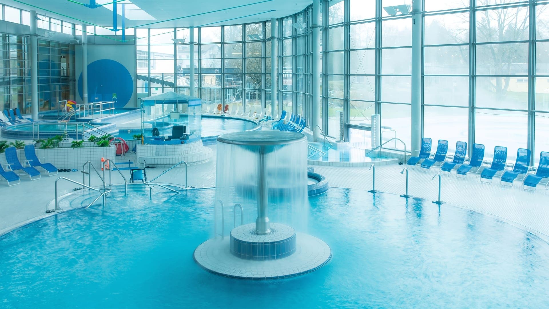 Indoor pool hall with a mushroom fountain and blue loungers by tall windows