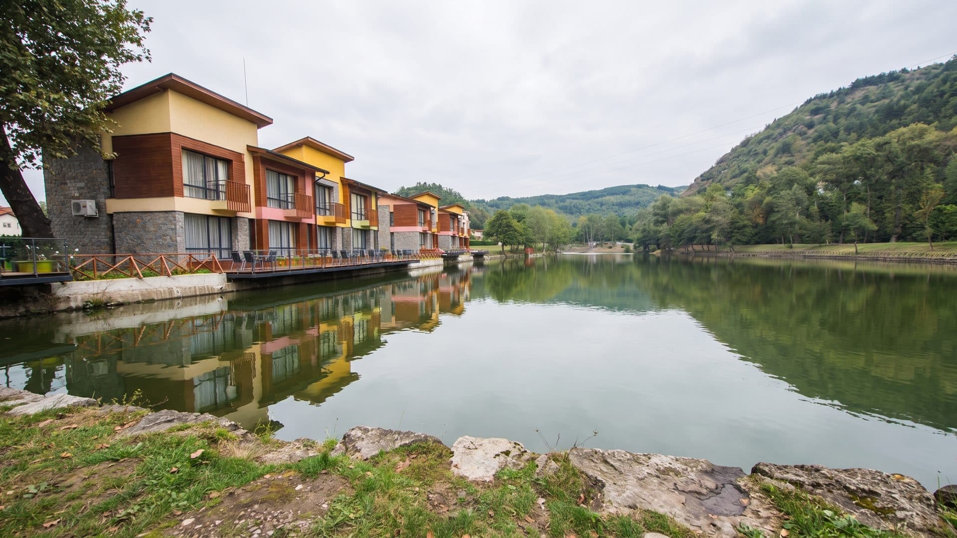 The balconied wing of the Waterside complex at Ognyanovo seen along the still water from the far bank, with the hills behind