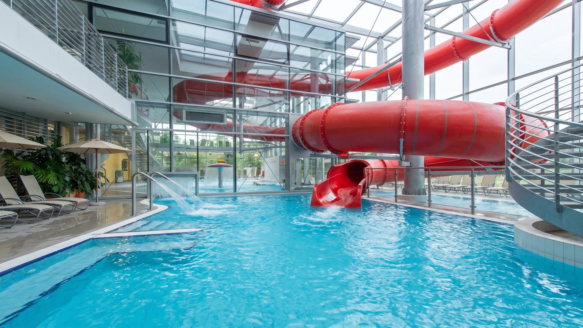 The indoor pool at Kurim under a glazed roof, with a red tube slide curling down to the water