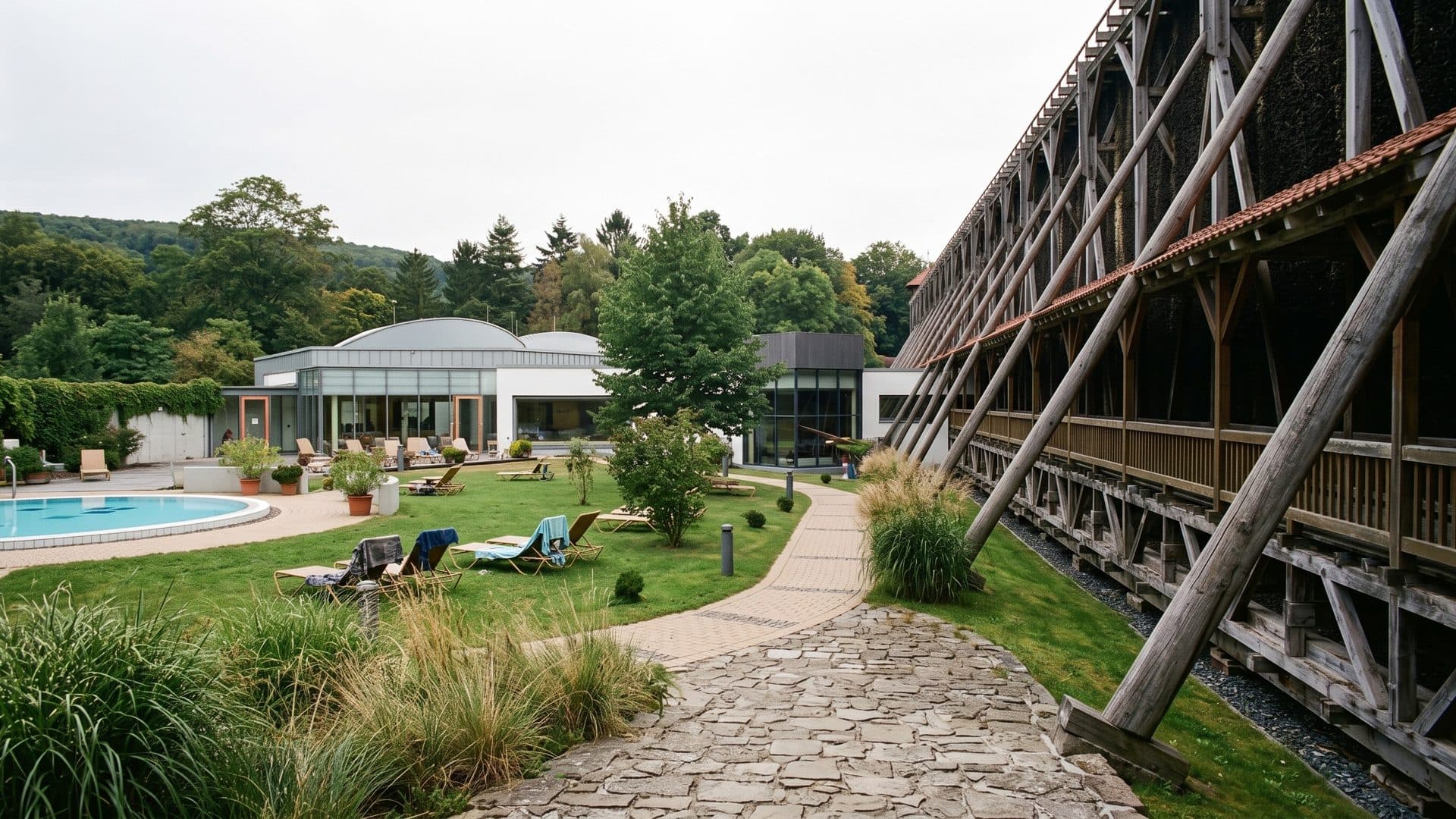 The lawn of the Werratal Therme between the timber graduation tower and the low round bath building, a small outdoor pool and loungers on the grass
