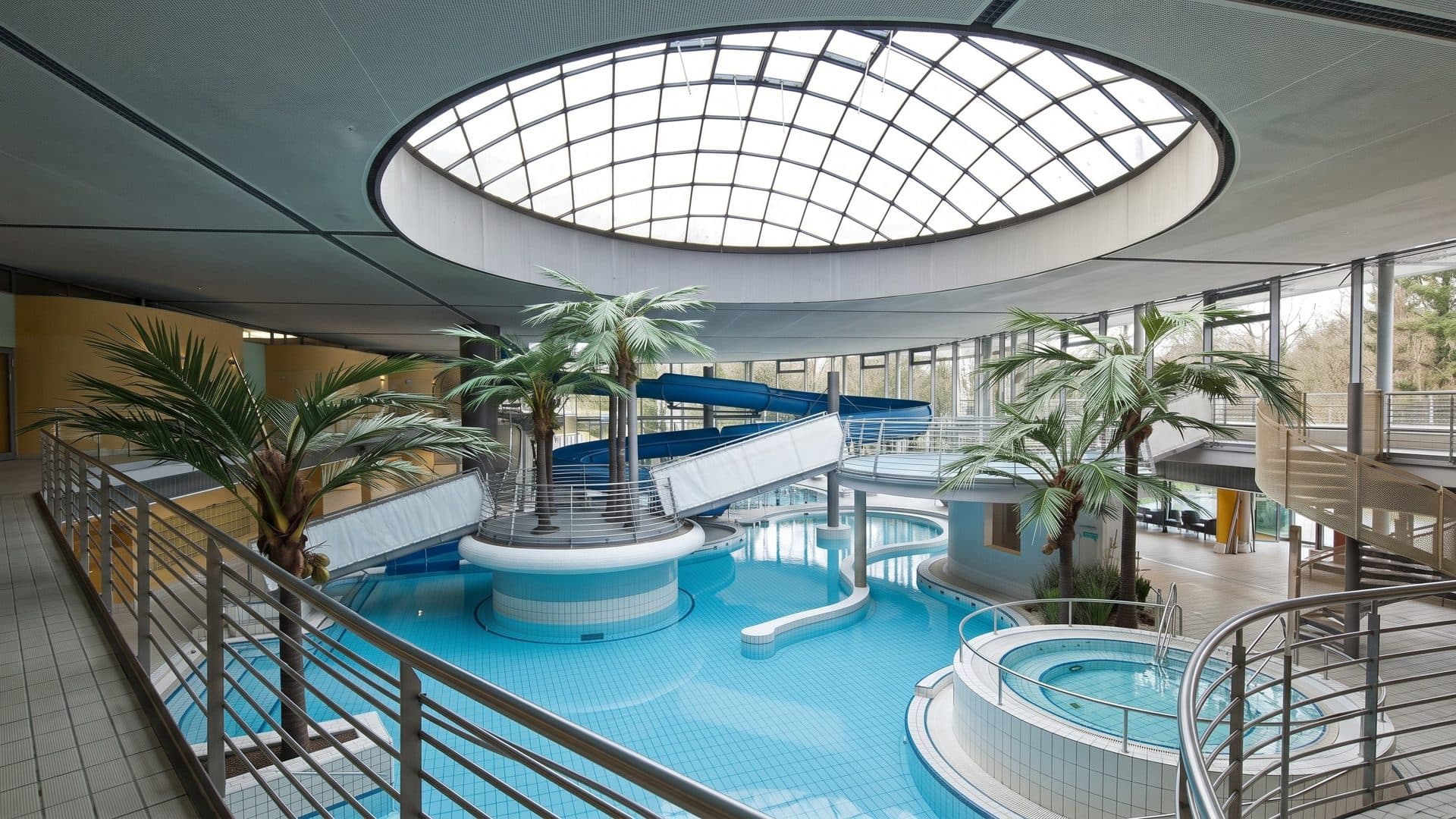 The leisure pool of the Westbad under a round glass skylight, palms on a central island, a white slide and a round whirlpool below the gallery