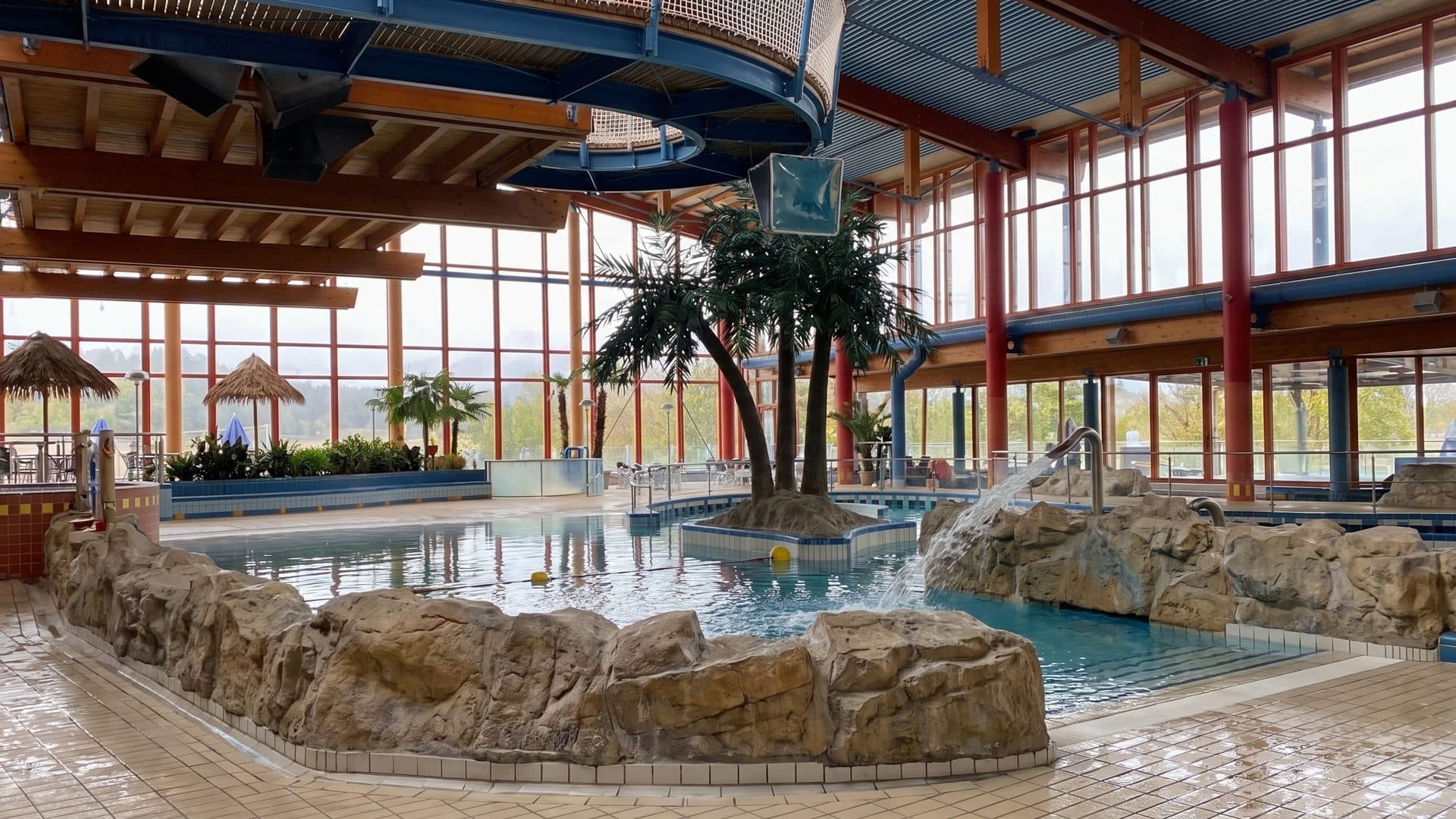 Indoor leisure pool with artificial rock edges and a palm island, under a red-framed glazed wall