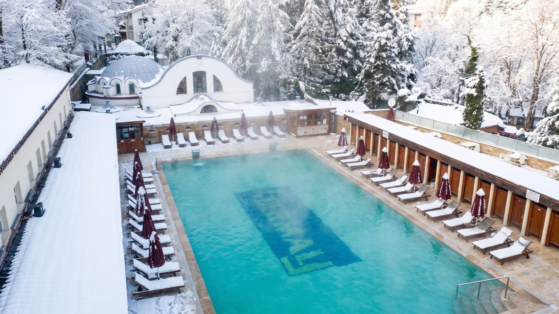 Steaming outdoor pool in winter, snow on the pines and cabins around it and a historic domed bathhouse behind.