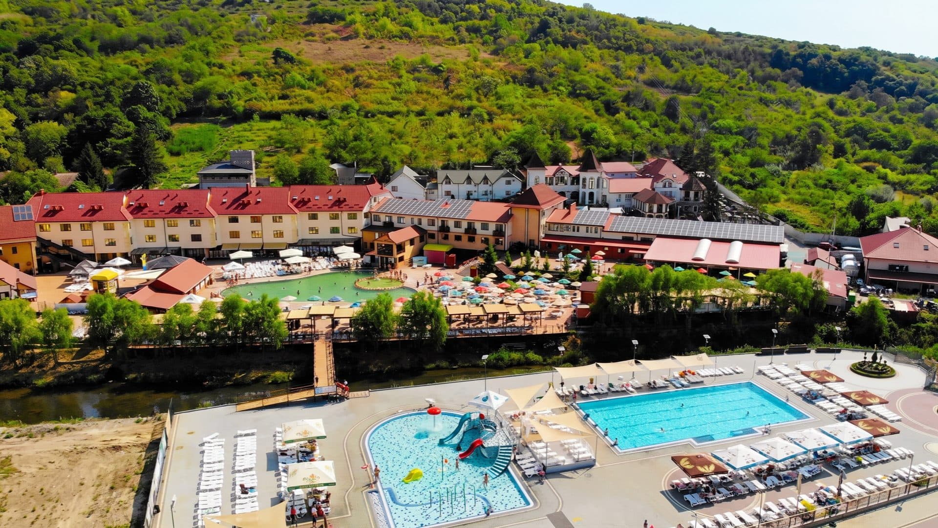 Aerial view of the thermal resort against a green hillside, with a childrens pool, a lap pool and a river running past.
