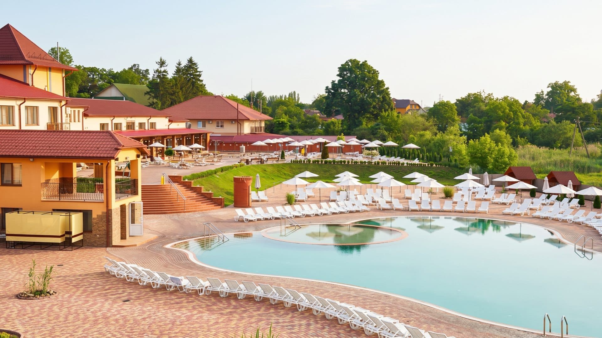 Curving outdoor thermal pool at a resort with orange-roofed lodges, rows of white loungers and parasols on a brick terrace.