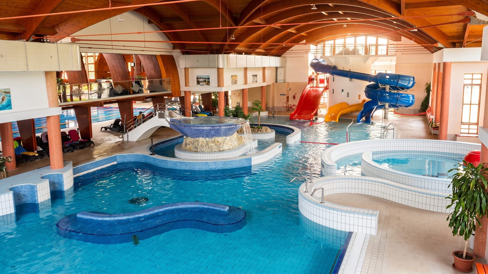 The indoor hall at Zsory Gyogy- es Strandfurdo in Mezokovesd, with curved pools, a waterfall feature and waterslides under a timber vaulted roof.