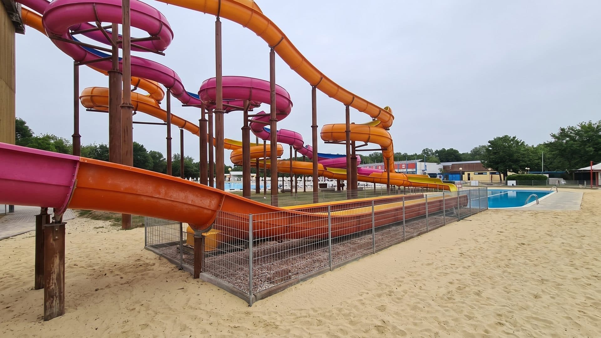 Cluster of orange and pink water slides on their timber tower above a sand-surrounded pool.