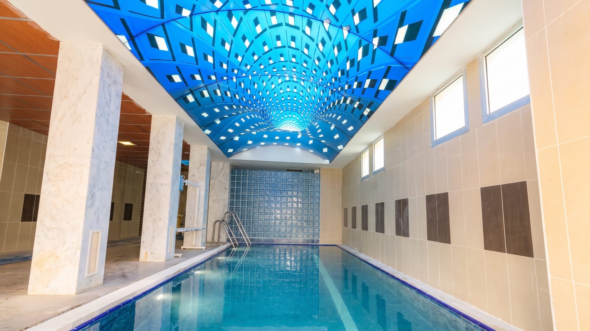 Indoor swimming pool beneath a vaulted blue backlit ceiling in a diamond pattern, flanked by marble columns and a glass-block wall.