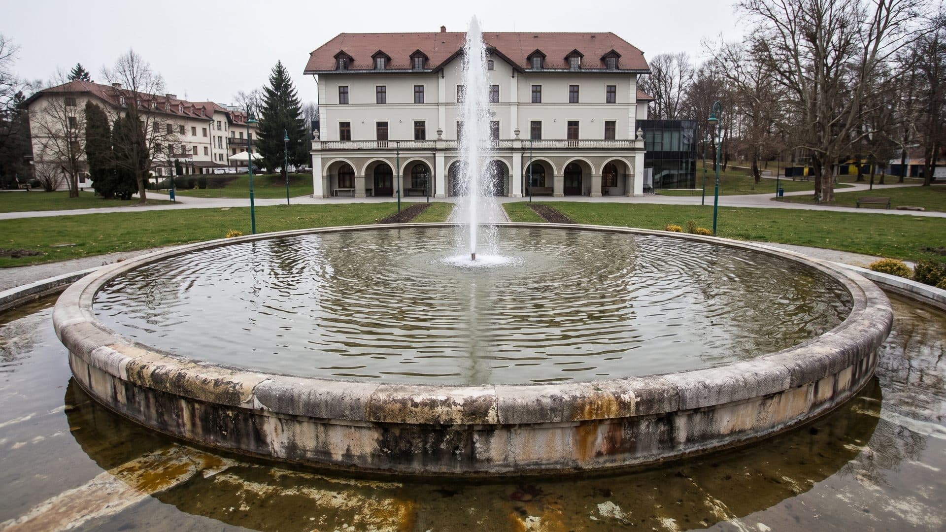 Round stone fountain basin in the park, the white spa house standing behind it among trees.
